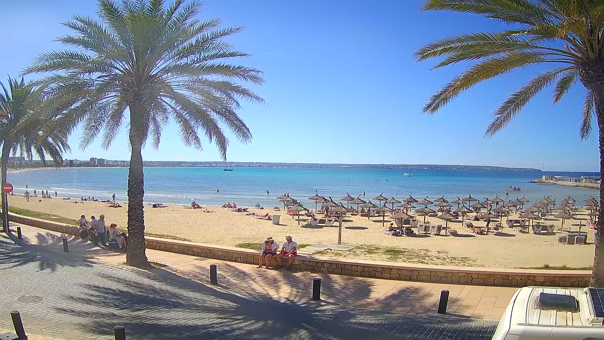 A sandy beach with numerous straw umbrellas and sunbathers is seen from a paved walkway, under a clear blue sky and bright sunshine, with calm turquoise water and palm trees in the foreground.