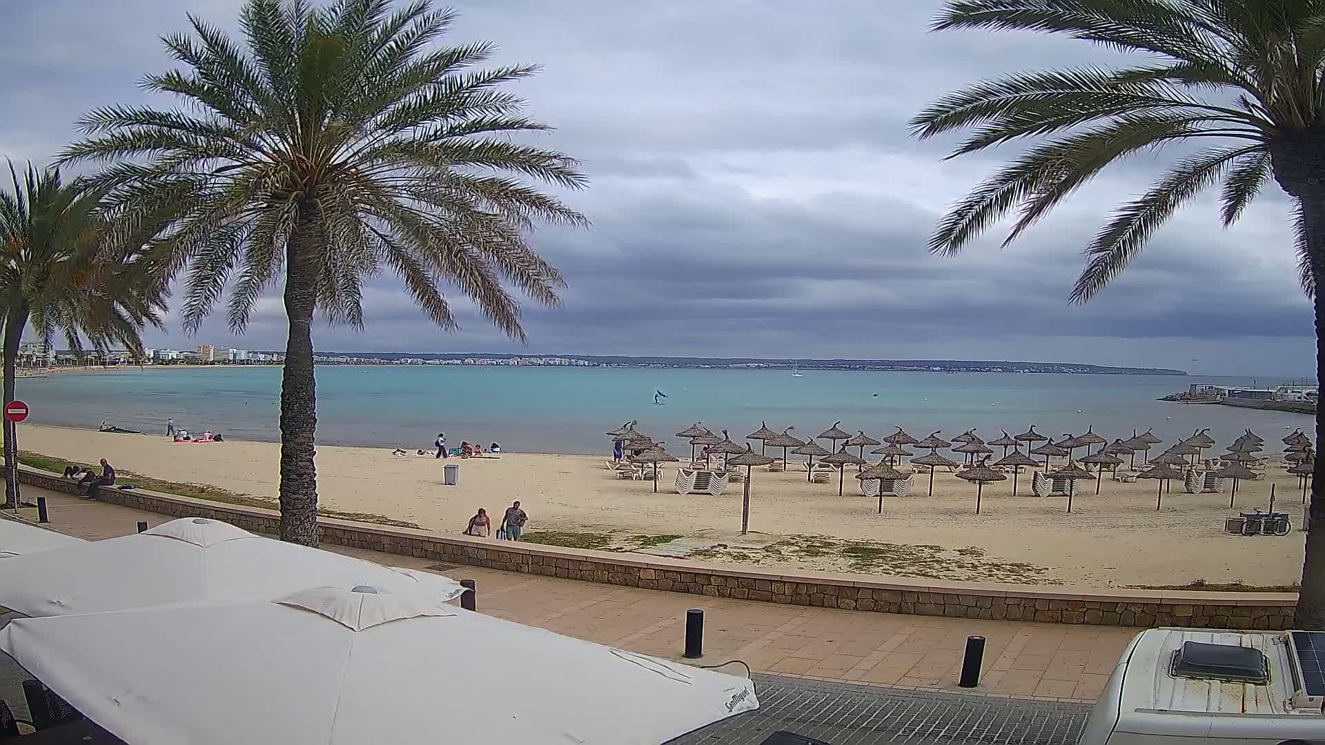 A sandy beach with numerous straw umbrellas, palm trees, and a few people is visible under a cloudy sky, overlooking a calm, turquoise body of water.