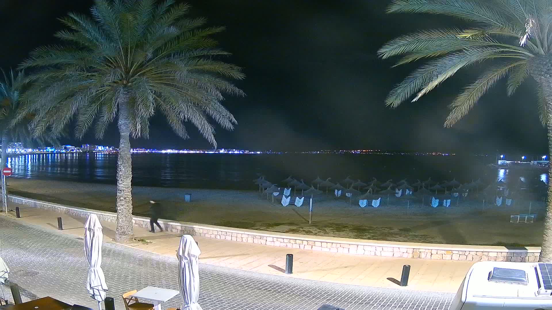 A nighttime beach scene shows a calm body of water with a city skyline in the distance, palm trees lining a paved walkway next to a sandy beach with closed umbrellas, and a person walking along the shore.