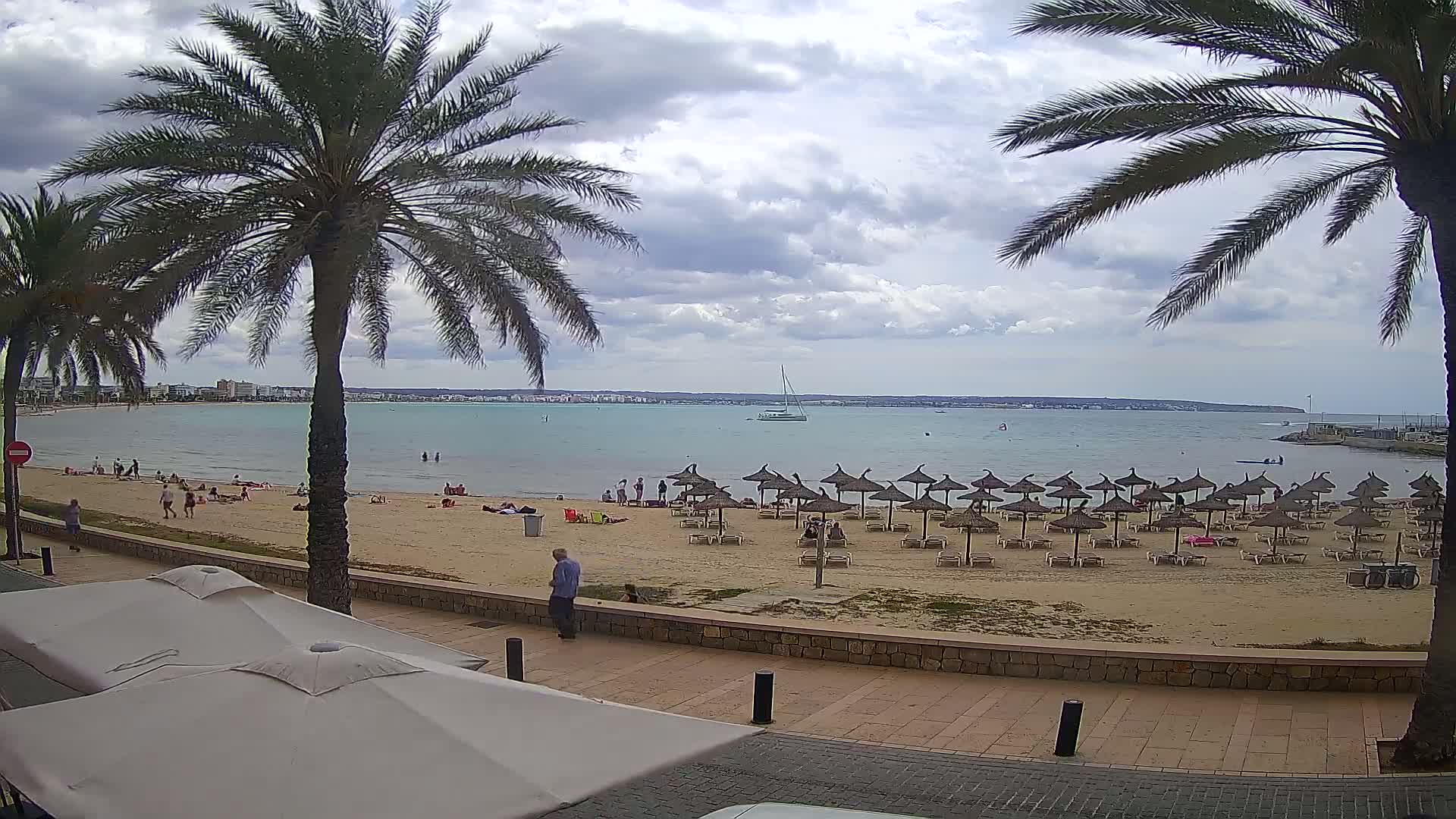 A sandy beach with many thatched umbrellas and scattered people is seen from a beachfront promenade under a cloudy sky, with calm water and a distant shoreline visible.