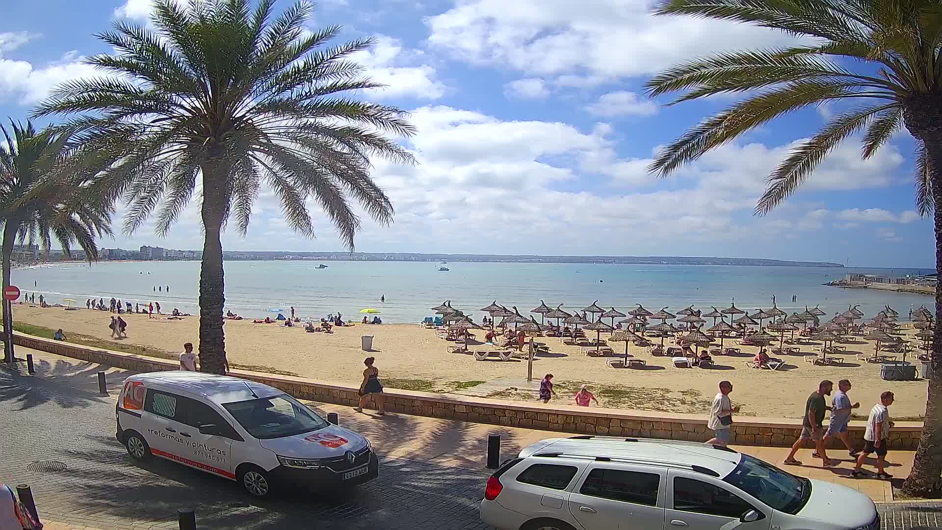 A sunny beach scene with palm trees, people relaxing under umbrellas, and several people walking along a paved walkway next to the sand.