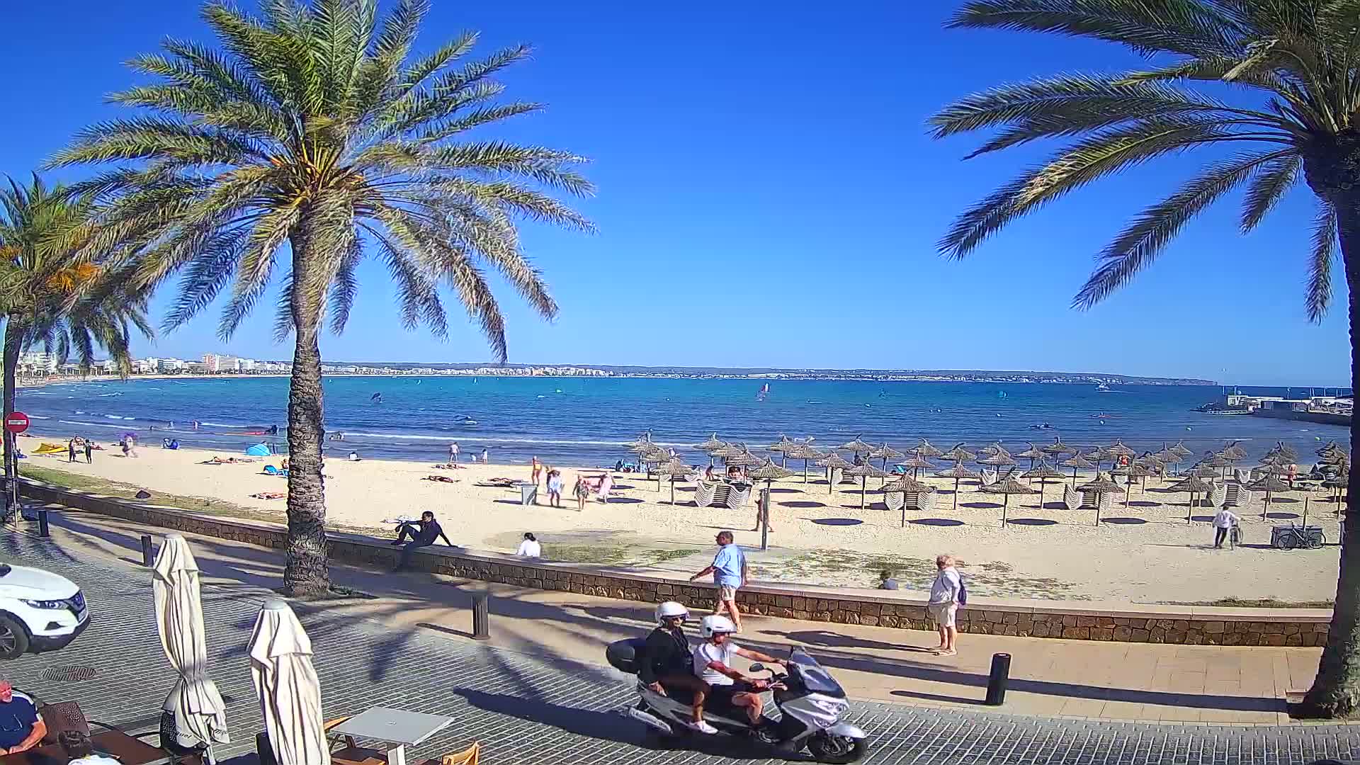 A sunny day at a sandy beach with calm blue water, palm trees, beach umbrellas, and people enjoying the outdoors.