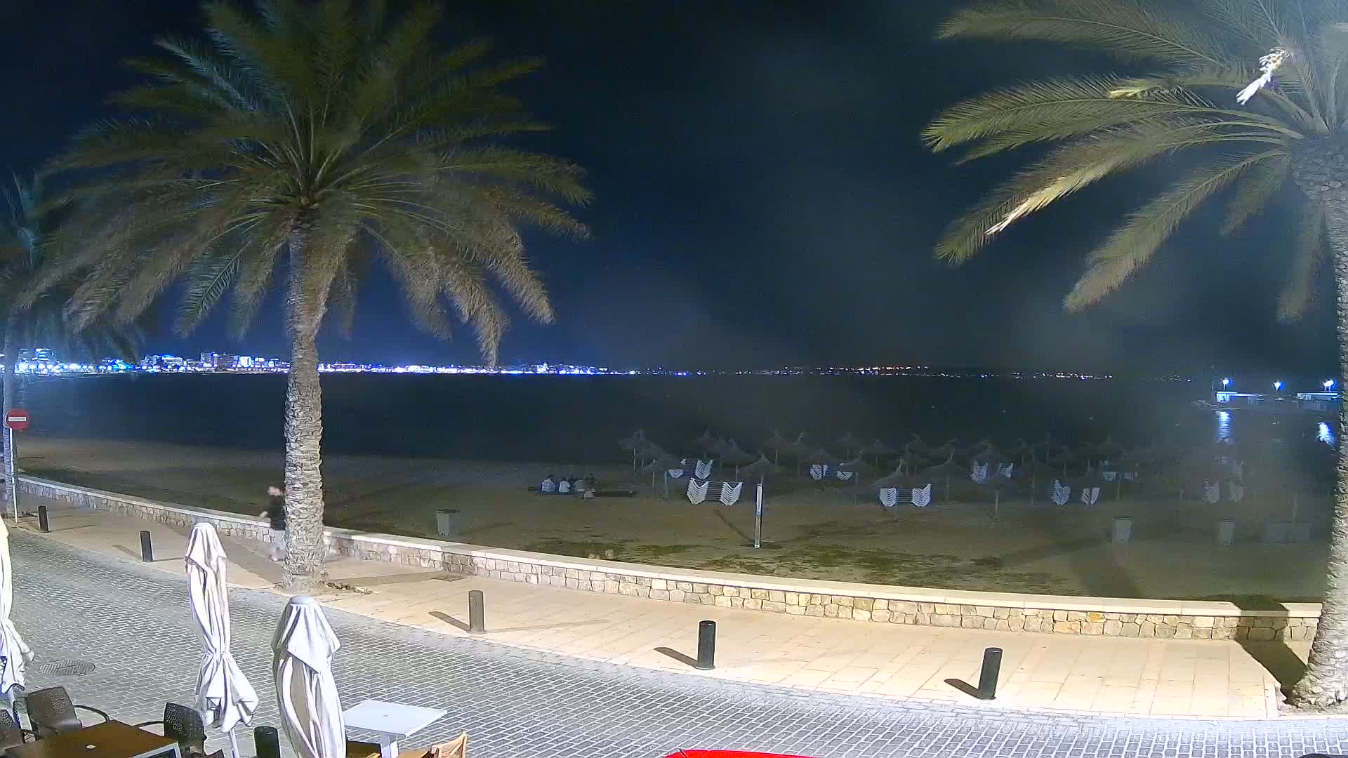 At night, a mostly empty beach with closed umbrellas is seen from a seaside promenade, with palm trees, a calm sea, and a city skyline faintly illuminated in the distance.