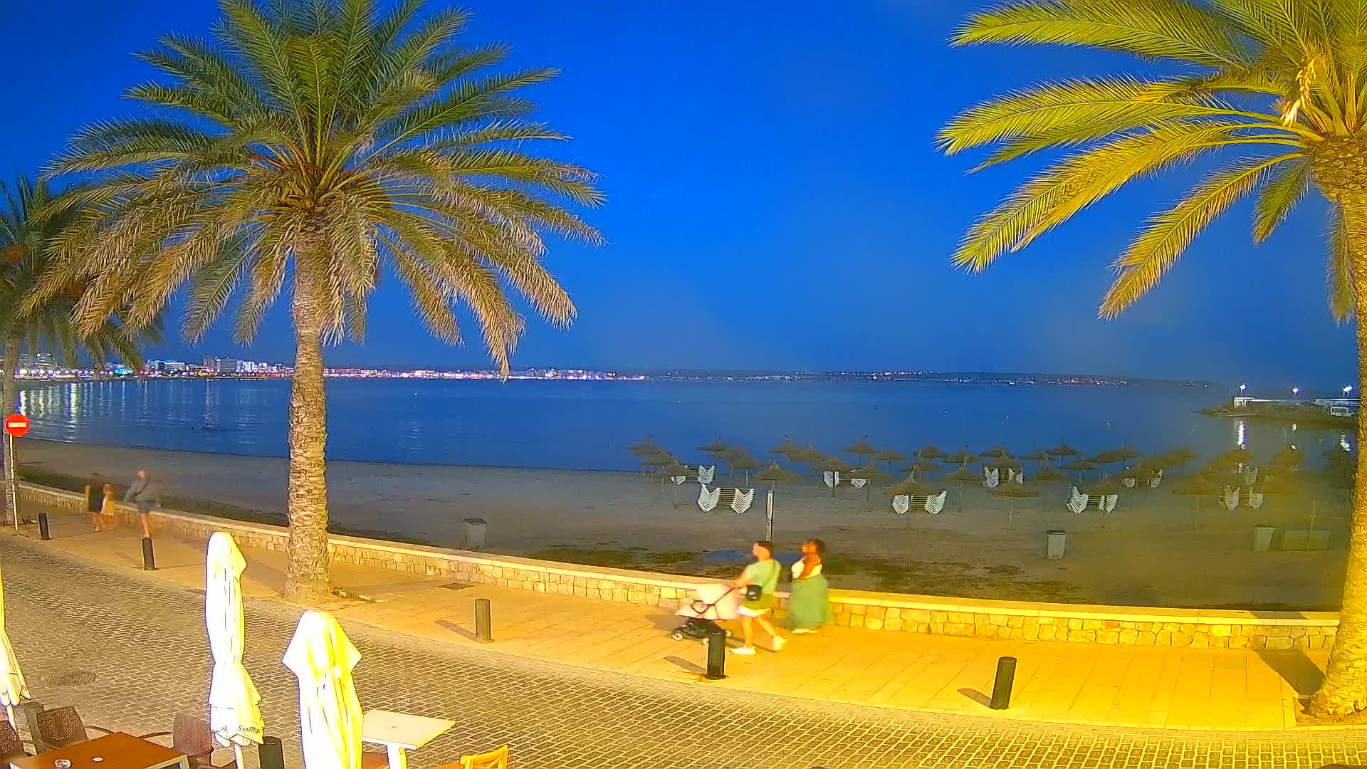 A beach at dusk with palm trees lining a paved walkway, several people walking, and numerous beach umbrellas on the sand.