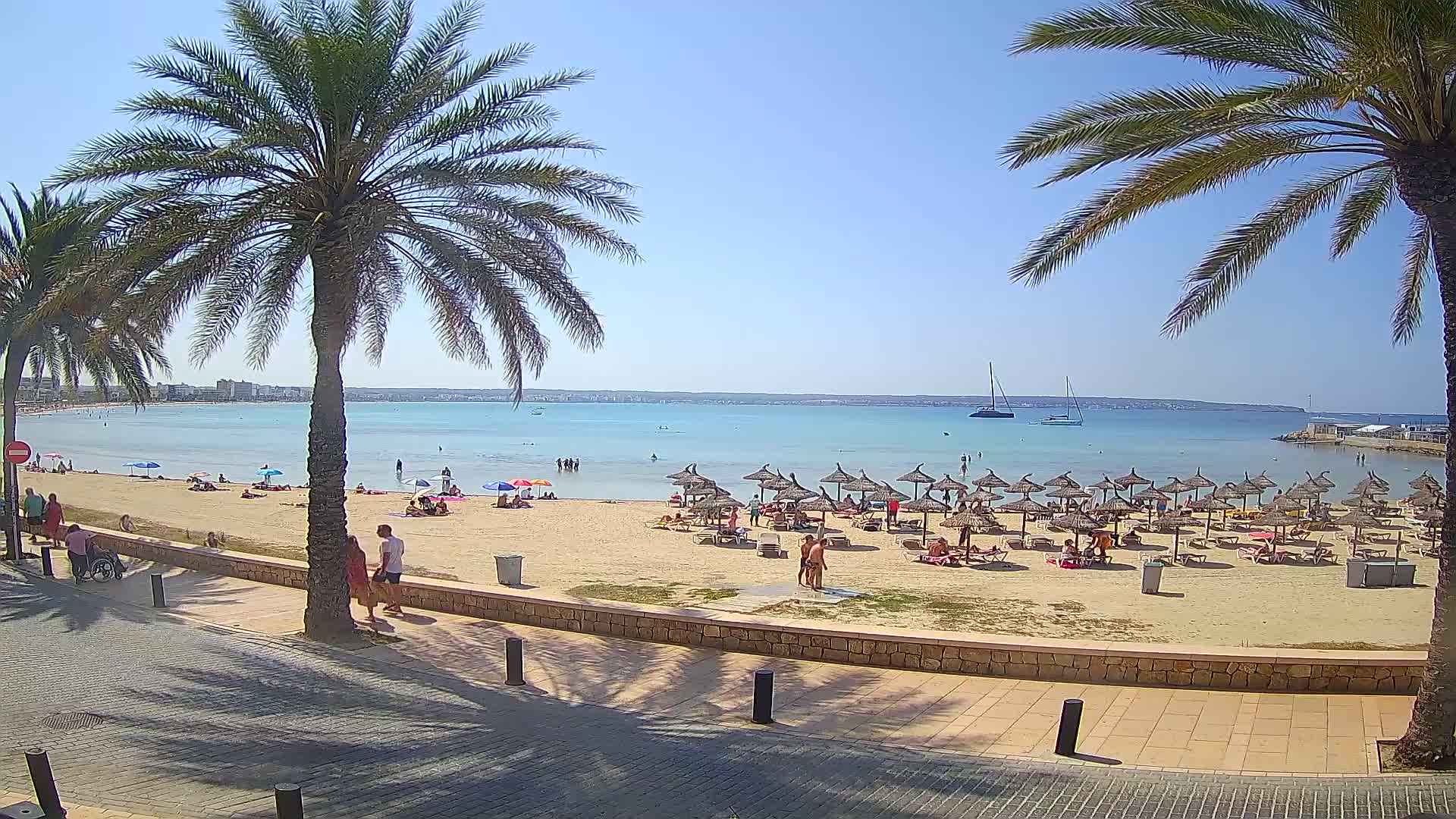 A sandy beach with numerous thatched umbrellas, people sunbathing and swimming in calm, clear turquoise water under a bright sunny sky, flanked by palm trees and a paved walkway.