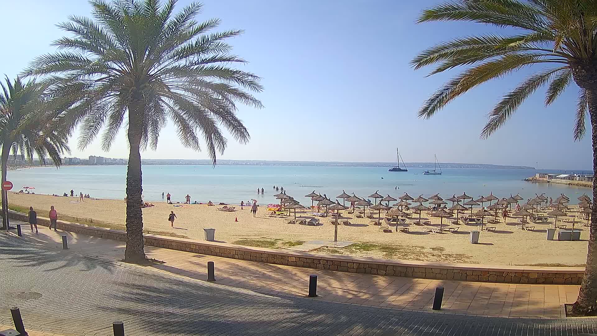 A sandy beach with numerous straw umbrellas and lounge chairs is shown, with calm, light-blue water and several people enjoying the sunny day.