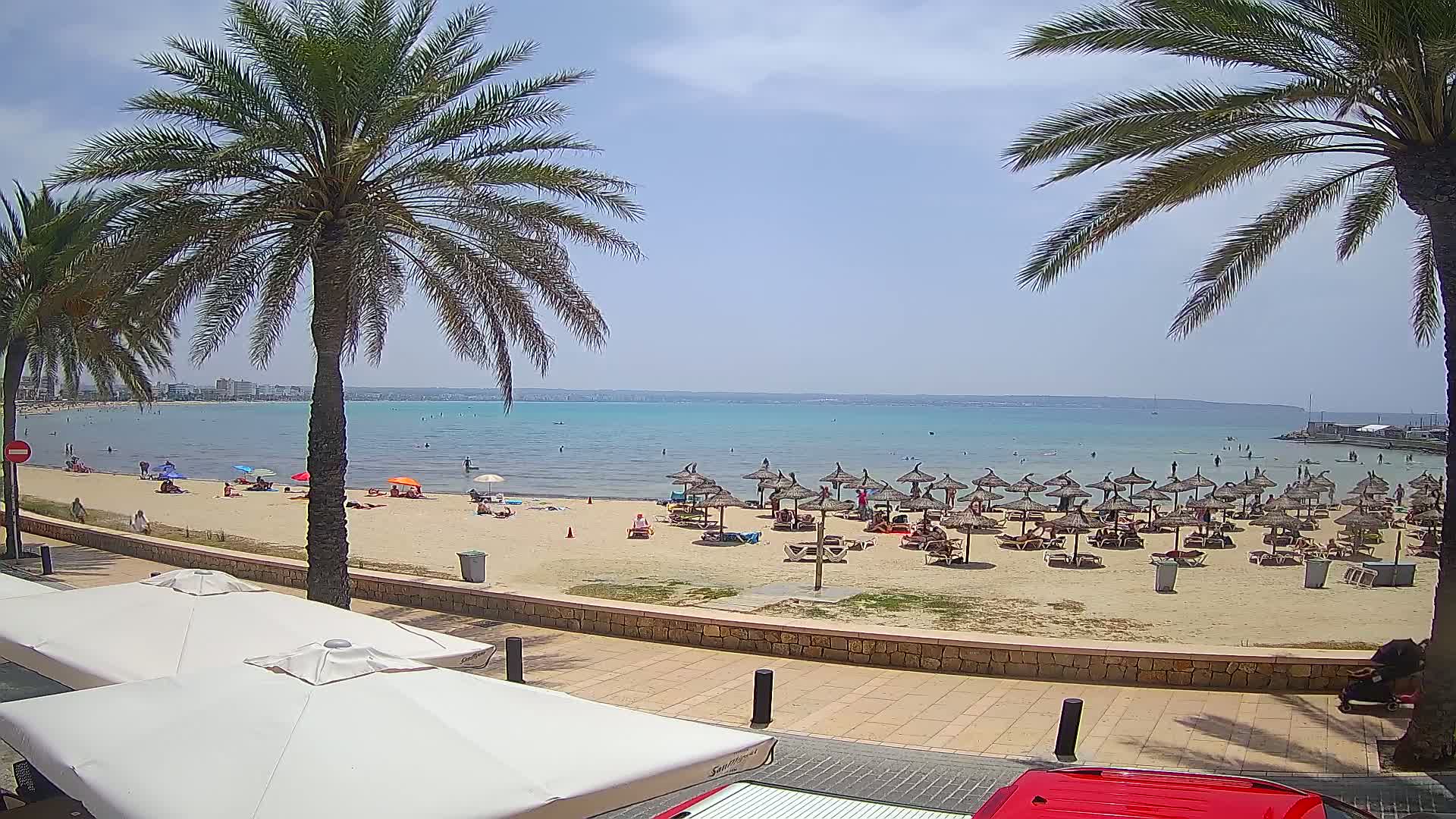 A sandy beach with numerous thatched umbrellas and sunbathers is visible from a promenade on a sunny day, with calm turquoise water extending to a distant shoreline.