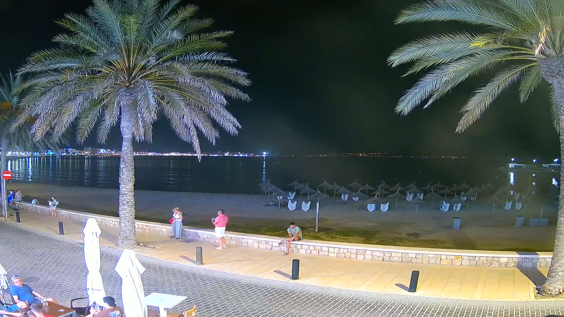 A sunny beach scene shows calm, turquoise water, many people enjoying the sand and shallows under numerous straw umbrellas, and palm trees lining a paved walkway.