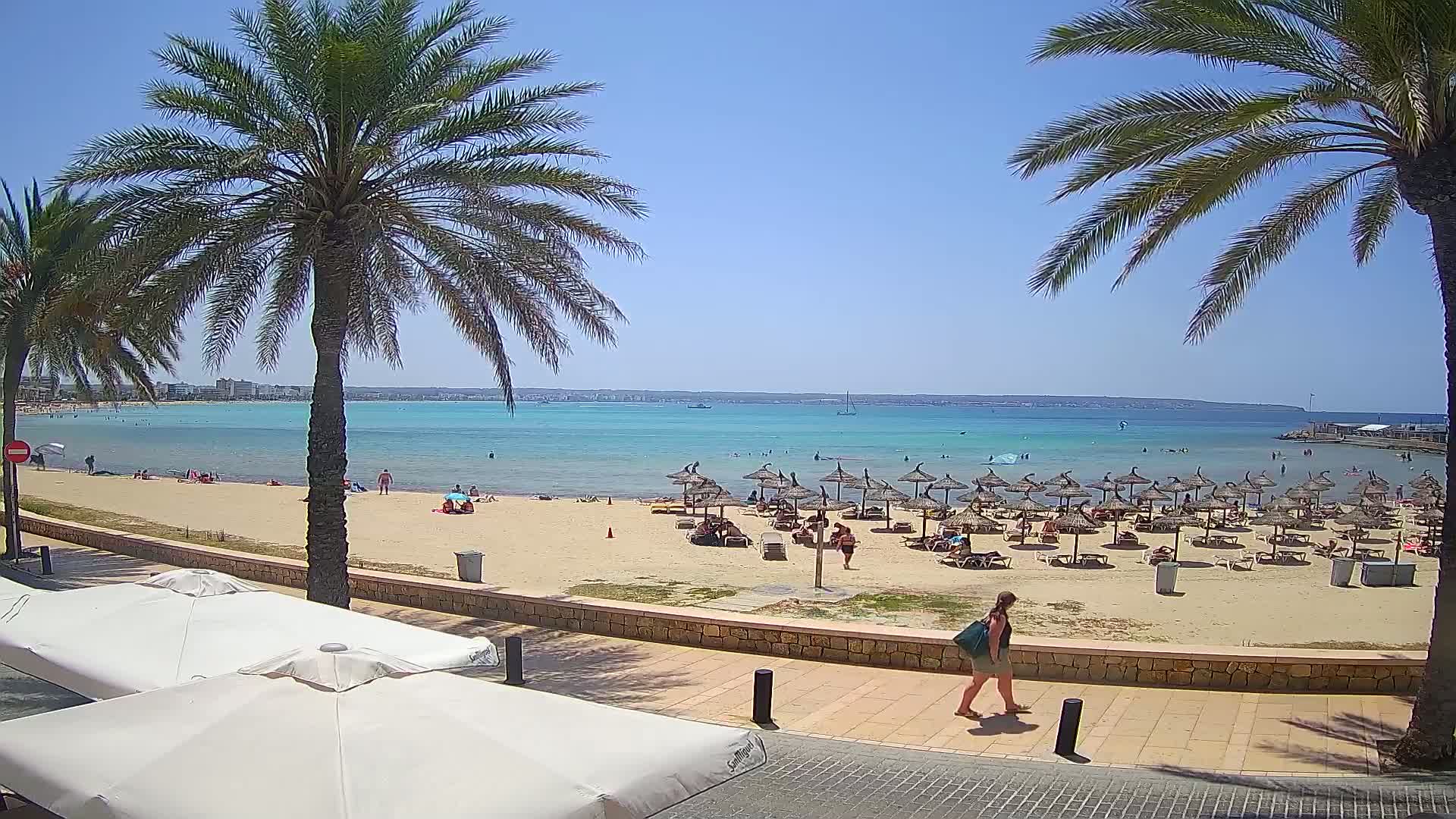 A sandy beach with numerous thatched umbrellas and sunbathers is seen from a boardwalk, overlooking calm, turquoise water under a clear, sunny sky.