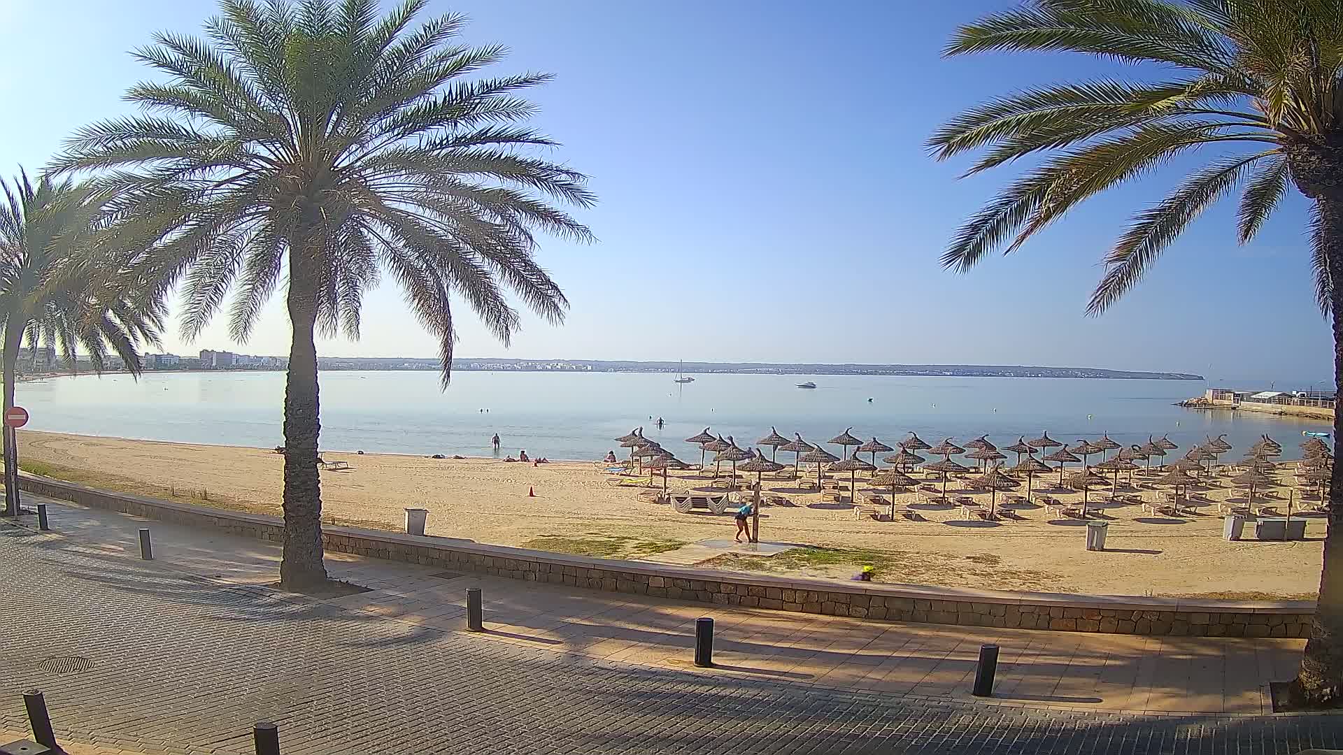 A sandy beach with numerous straw umbrellas and lounge chairs is situated beside a calm, clear body of water under a bright, sunny sky; palm trees line the paved walkway leading to the beach.