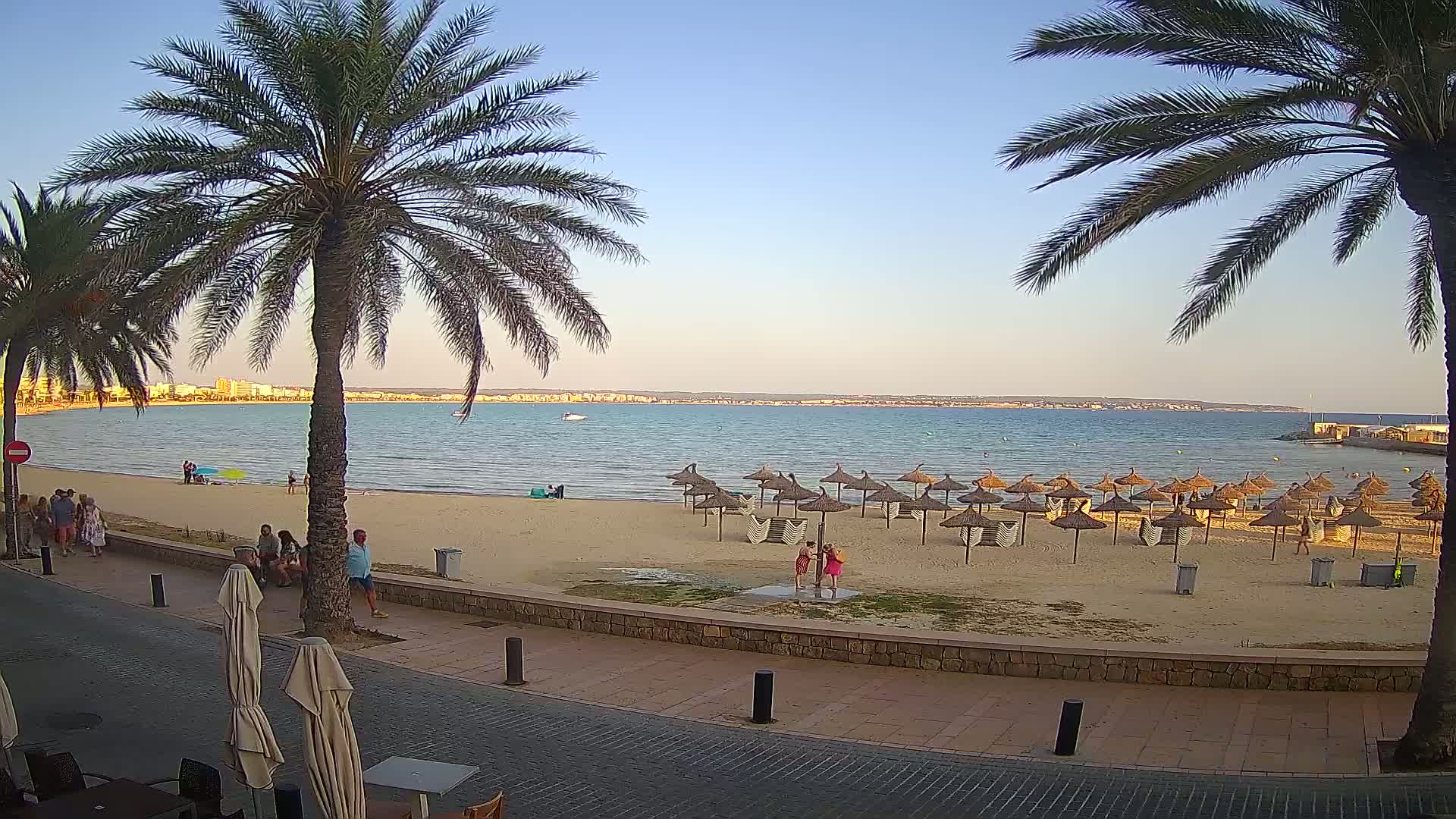 A sandy beach with numerous thatched umbrellas, palm trees, and several people is visible from a paved walkway, all under a clear, calm evening sky.
