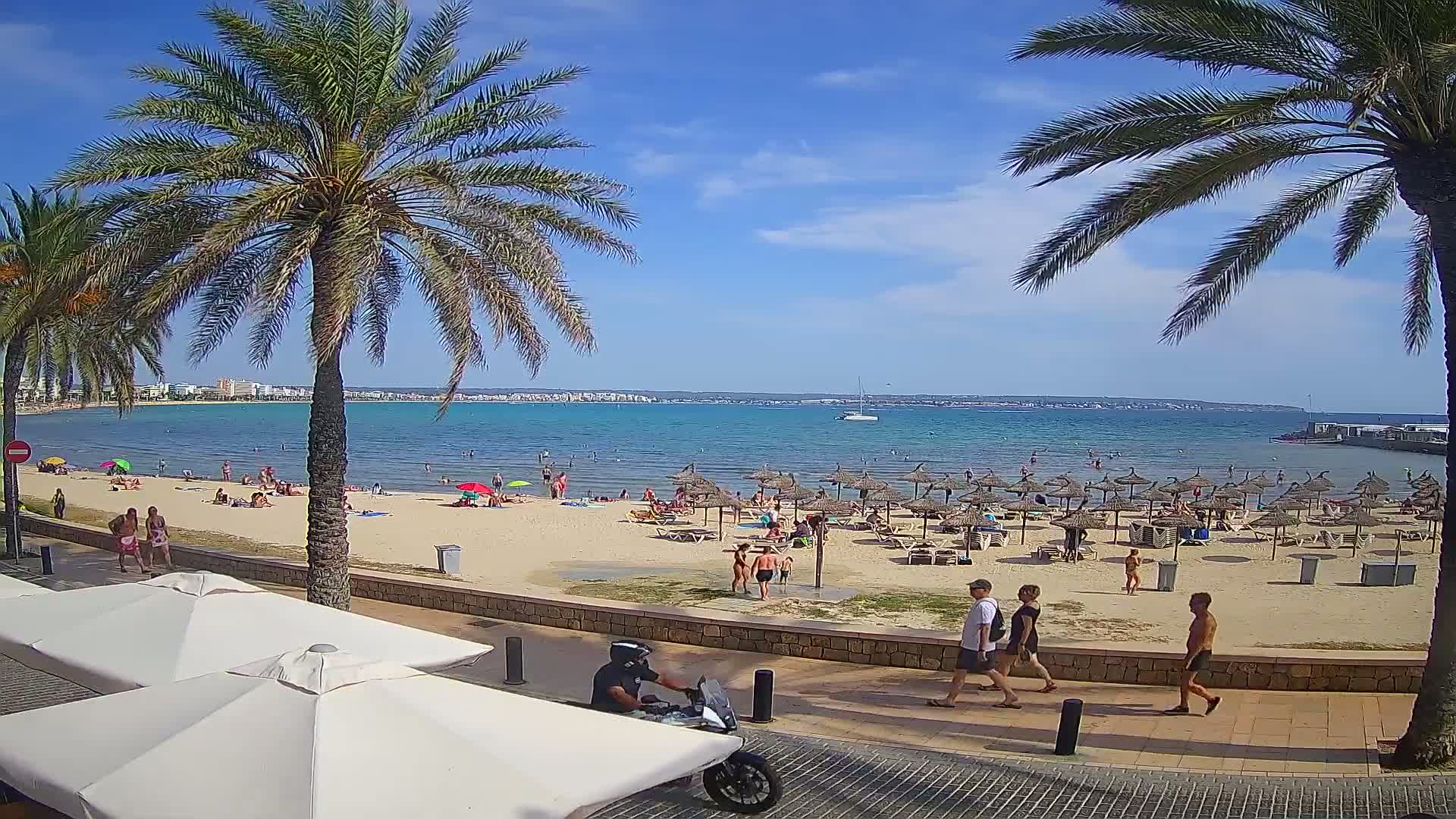 A sunny beach scene shows a sandy shore with numerous beach umbrellas and people swimming and sunbathing, all under a mostly clear blue sky.