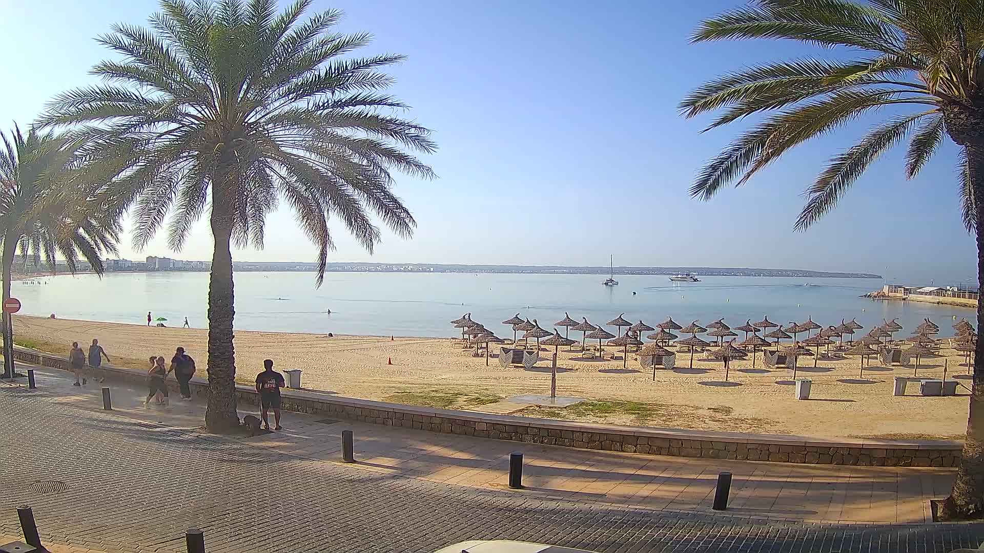A sandy beach with numerous straw umbrellas, several people walking along a paved walkway, and calm water under a clear blue sky.