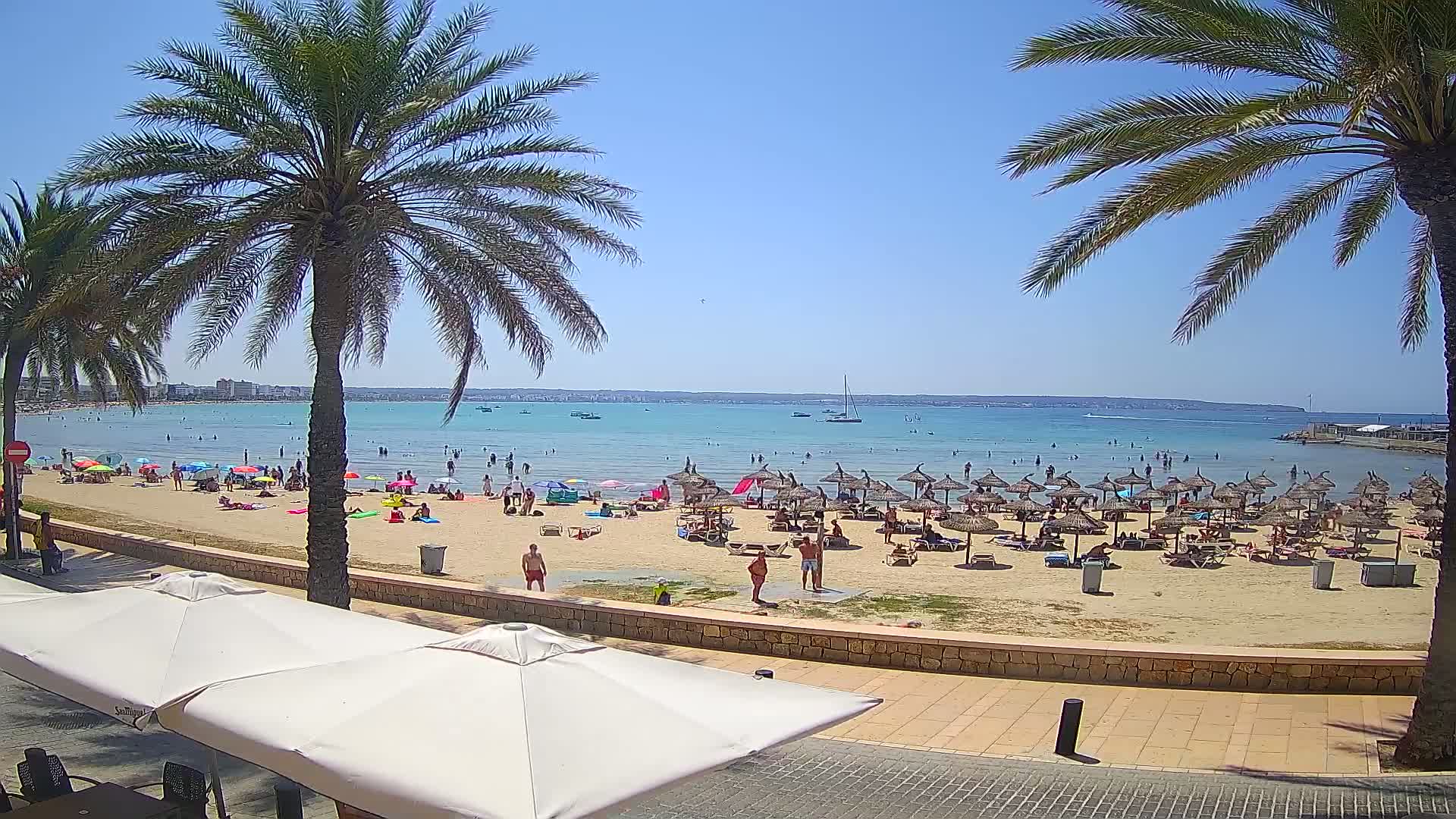 A sunny beach scene shows a sandy shore dotted with numerous beach umbrellas and people swimming and relaxing in shallow, calm, turquoise water, all under a clear blue sky.