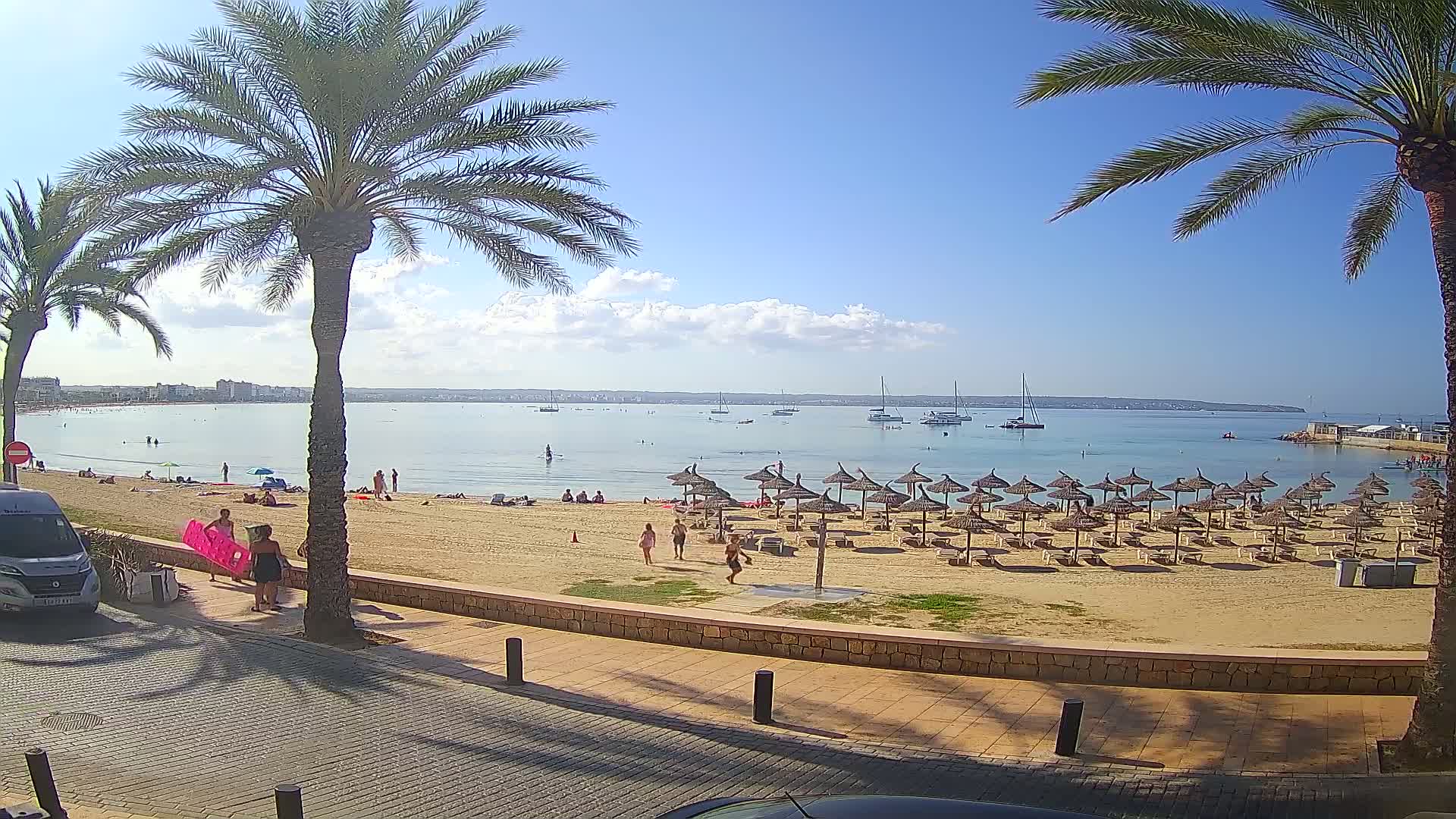 A sandy beach with numerous straw umbrellas and several people is seen from a slightly elevated vantage point, overlooking calm, clear water with boats in the distance under a mostly sunny sky.