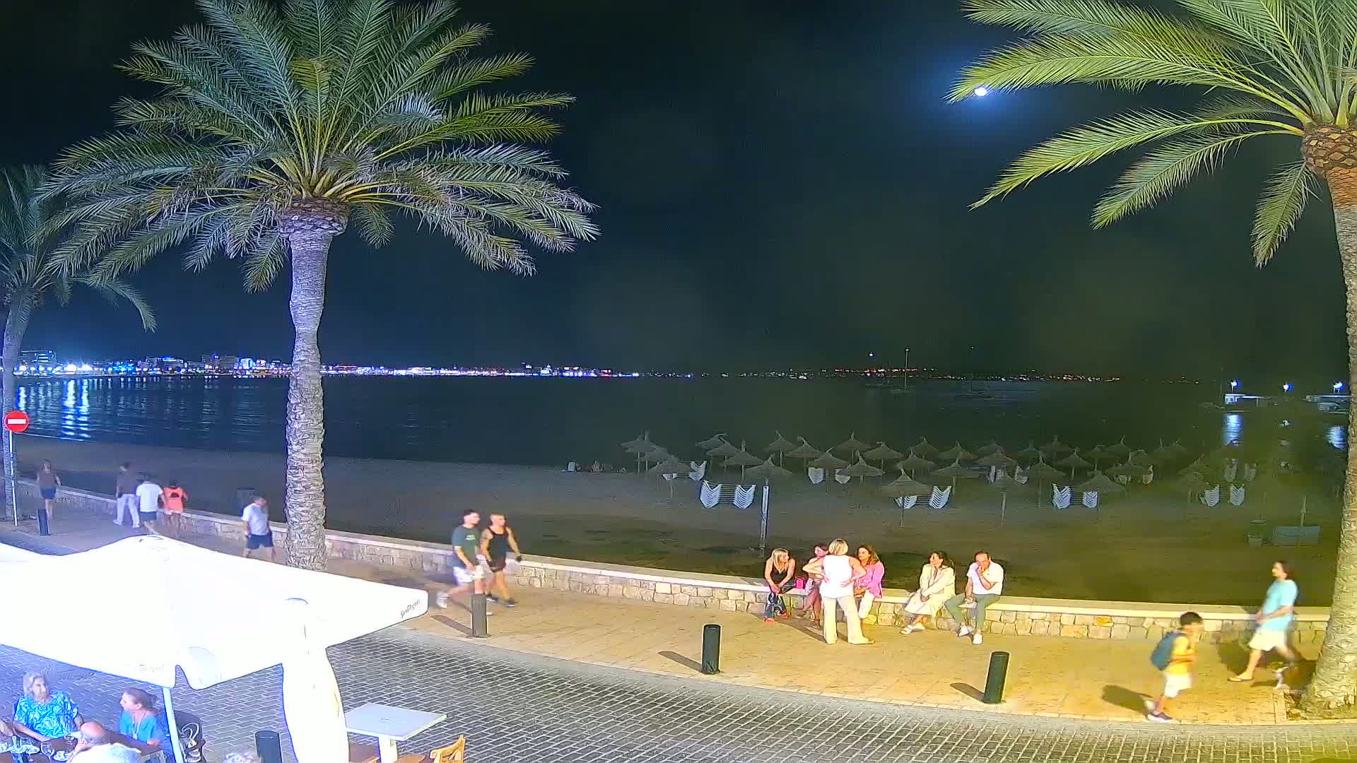 A nighttime beach scene shows people strolling along a paved walkway, some seated on a low wall overlooking a sandy beach with closed umbrellas, a calm body of water, and a city skyline in the distance under a moonlit sky.