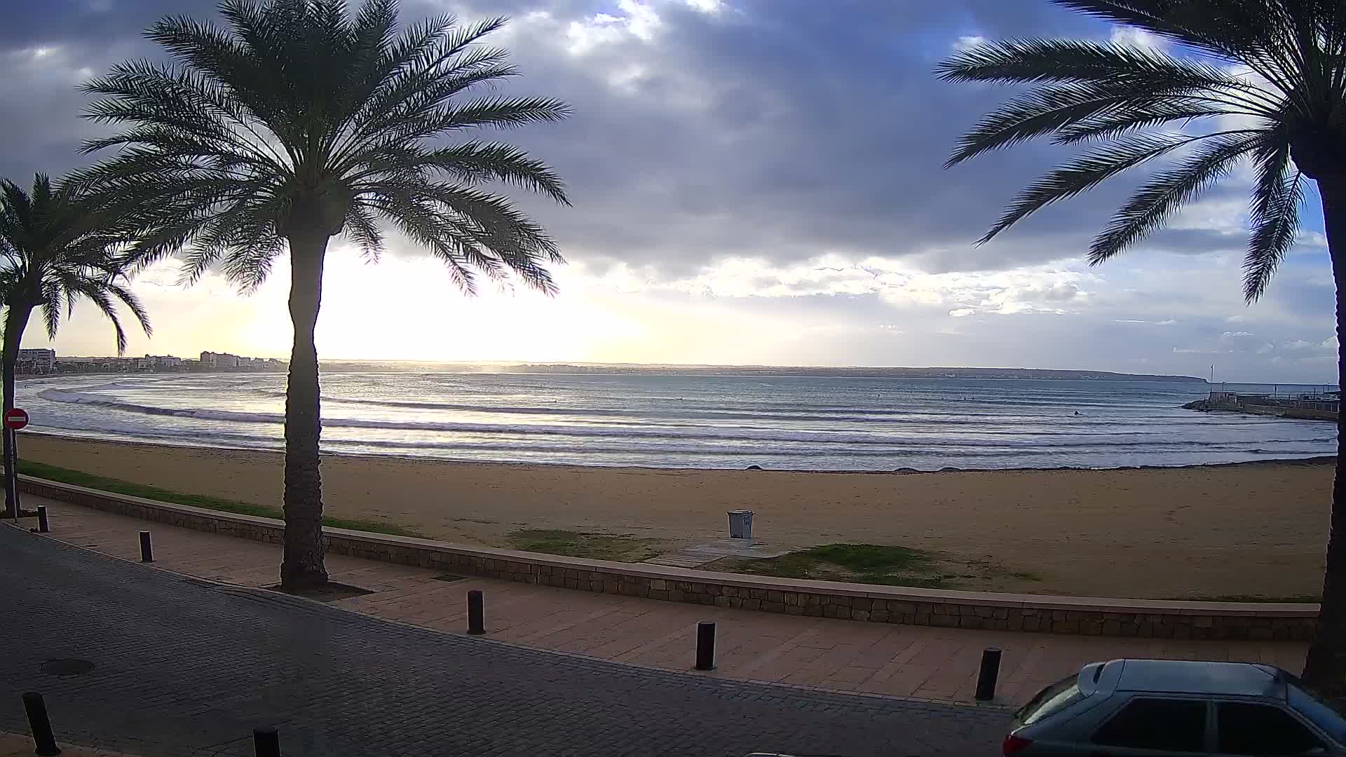 A vibrant tropical beach scene features palm trees framing a sandy shore and ocean under a dramatic, partly cloudy sky with bright sun, bordered by a paved promenade and a distant coastal town.