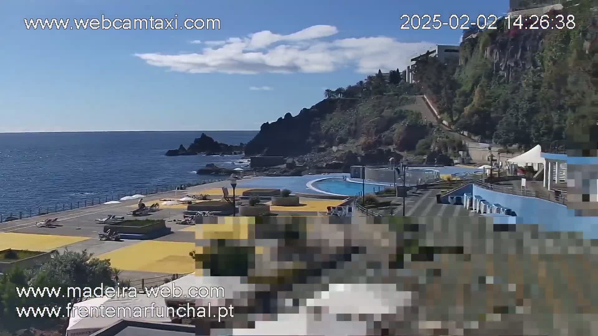 An outdoor scene shows a seaside pool area with a rocky cliff and ocean under a partly cloudy sky.