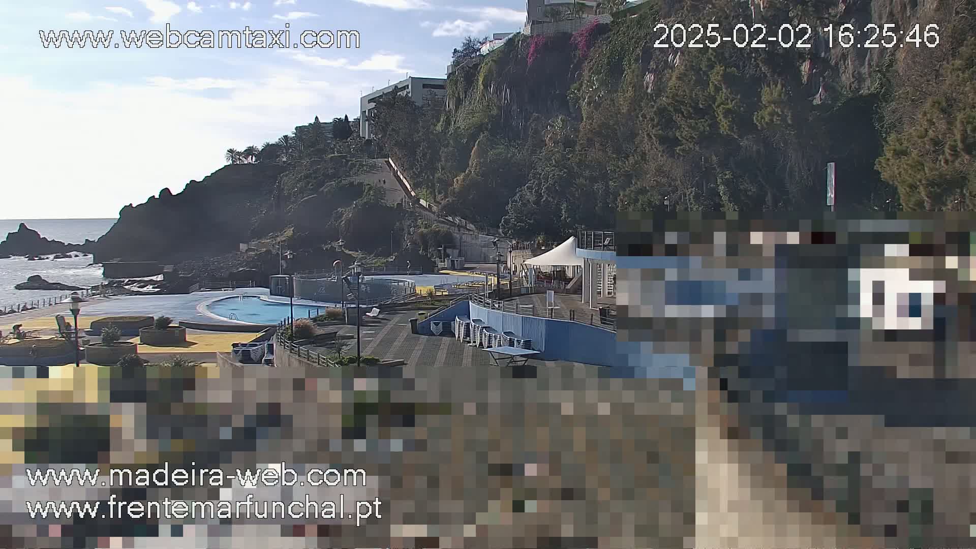 An outdoor scene on a sunny day shows an oceanside pool area with a cliffside backdrop, featuring buildings and lush green vegetation.