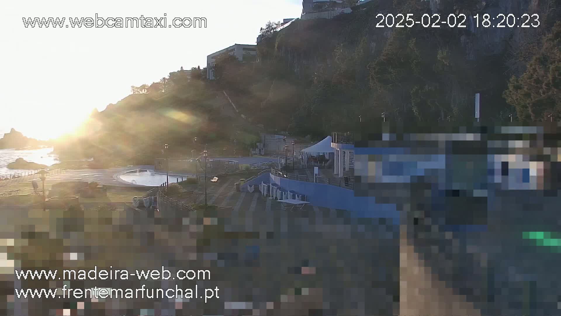 A coastal scene at sunset shows a swimming pool area, buildings, and a cliff overlooking the ocean.