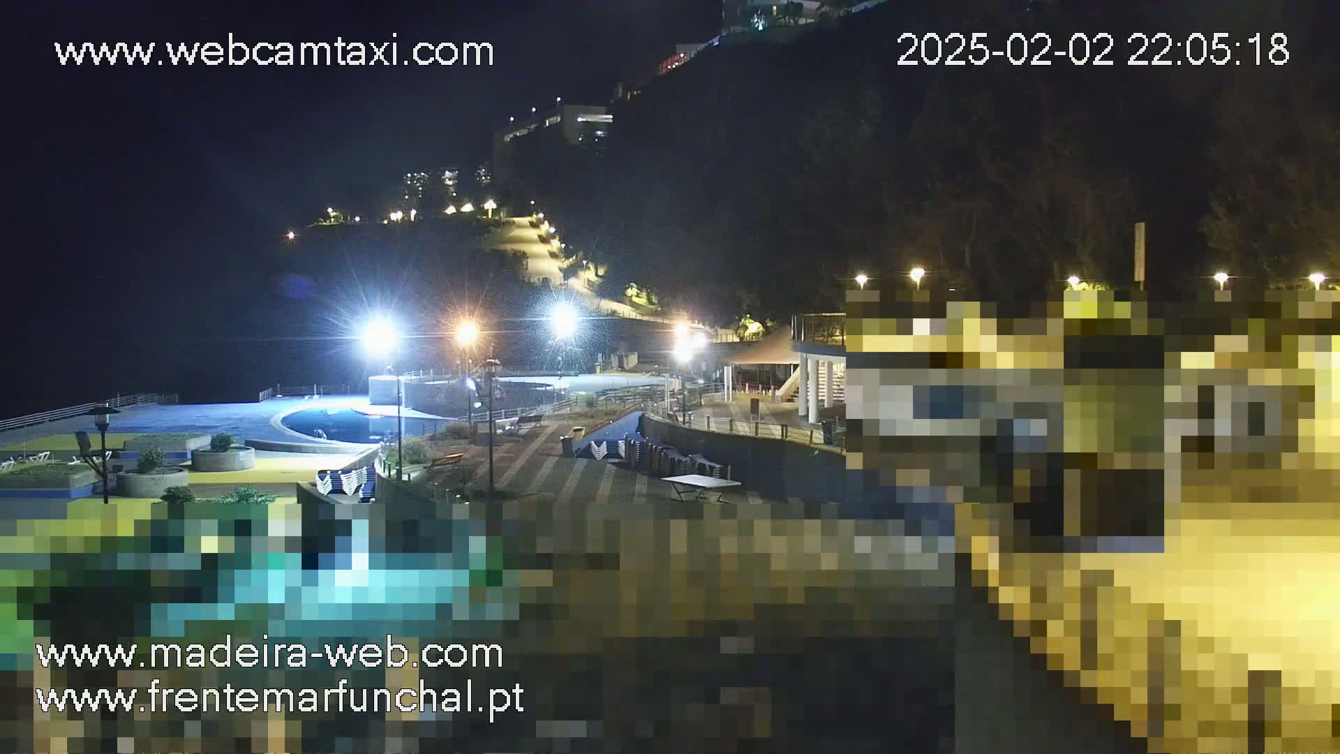 A nighttime view of a coastal area with a swimming pool, illuminated pathways, and buildings on a hillside.