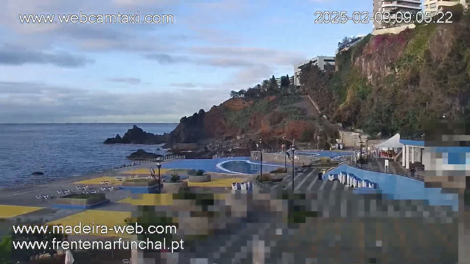 An outdoor scene on a partly cloudy day shows a seaside pool area with buildings on a cliff overlooking the ocean.