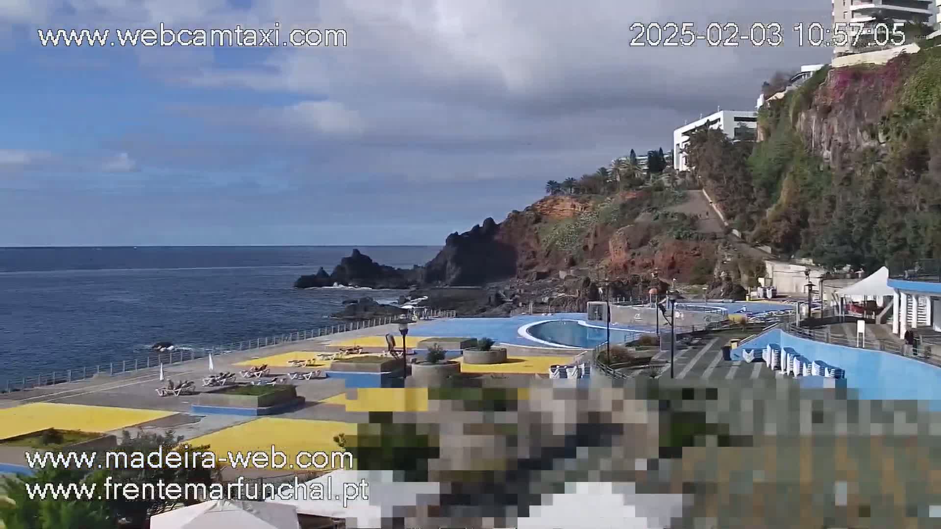 A seaside pool area with a curved pool and surrounding buildings sits on a rocky cliff overlooking a calm ocean under a partly cloudy sky.