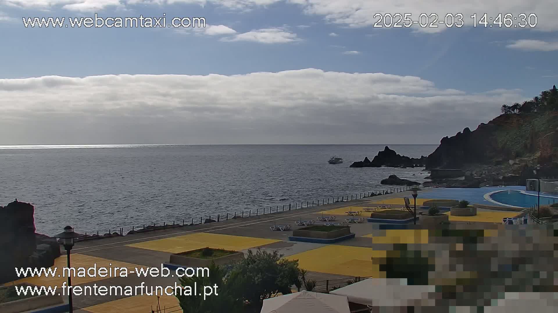 A partly cloudy day overlooks a calm ocean next to a paved waterfront area with a swimming pool and some lounge chairs.