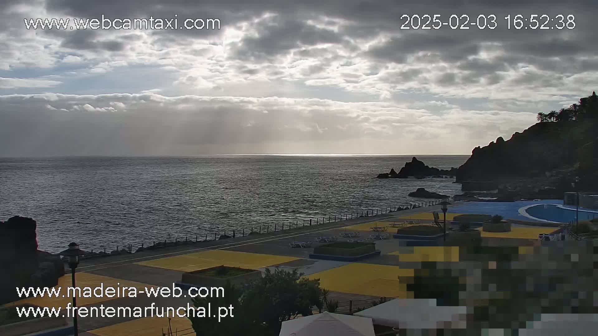 A mostly cloudy day overlooks a dark-blue ocean with dark rocky cliffs and a paved area with lounge chairs and a swimming pool.