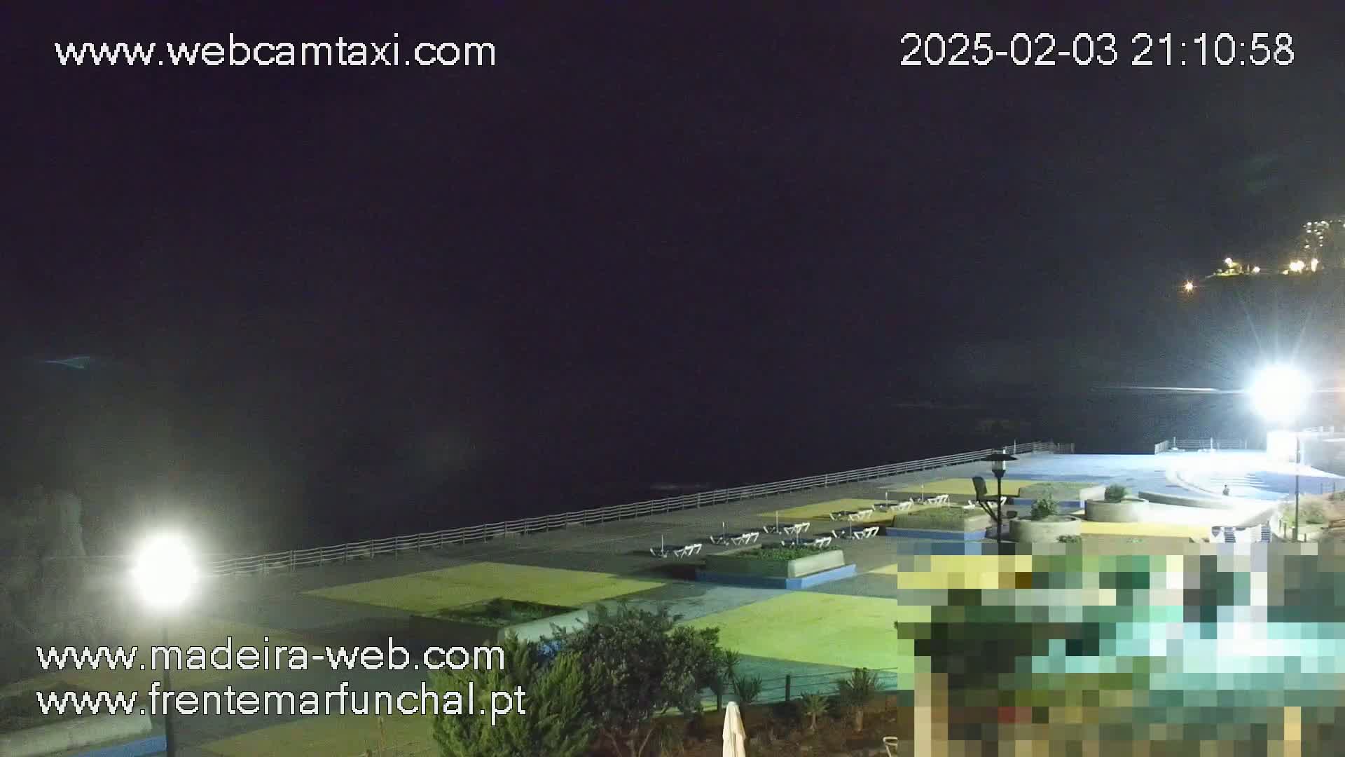 A nighttime view of a mostly empty paved seaside area with some lights, plants, and a few pieces of furniture.