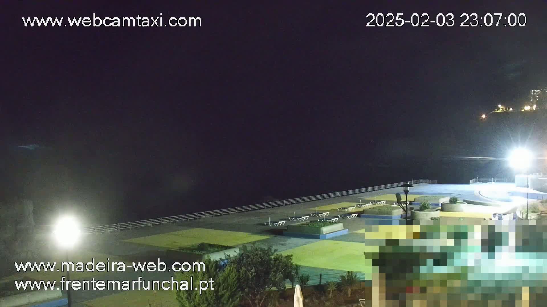A nighttime, seaside view shows an empty paved area with some landscaping, lights, and a few chairs near a dark ocean under a clear, dark sky.