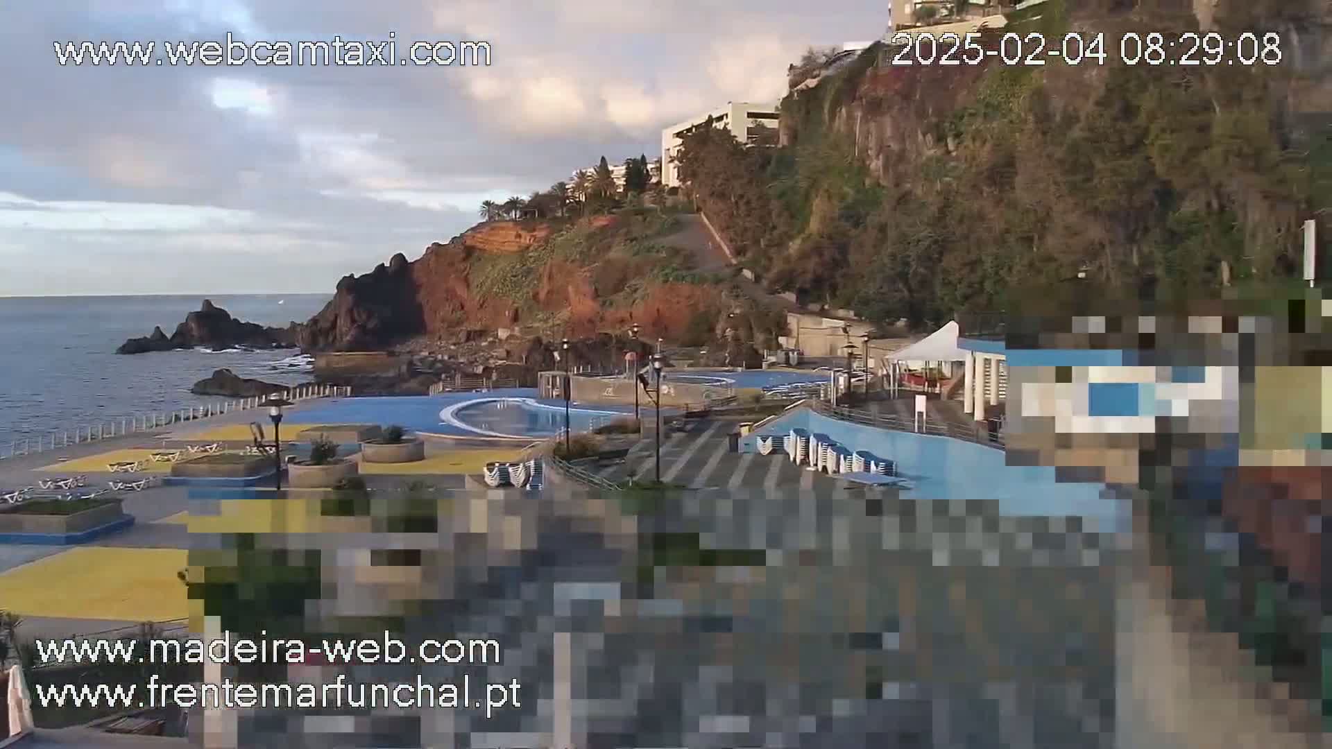 An outdoor seaside pool complex is situated on a rocky cliffside overlooking a calm ocean under a partly cloudy sky.