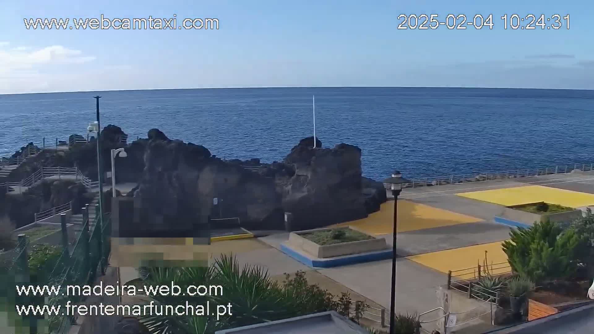A rocky outcrop juts into a calm, blue ocean under a clear sky, beside a paved area with yellow sections and low-lying landscaping.