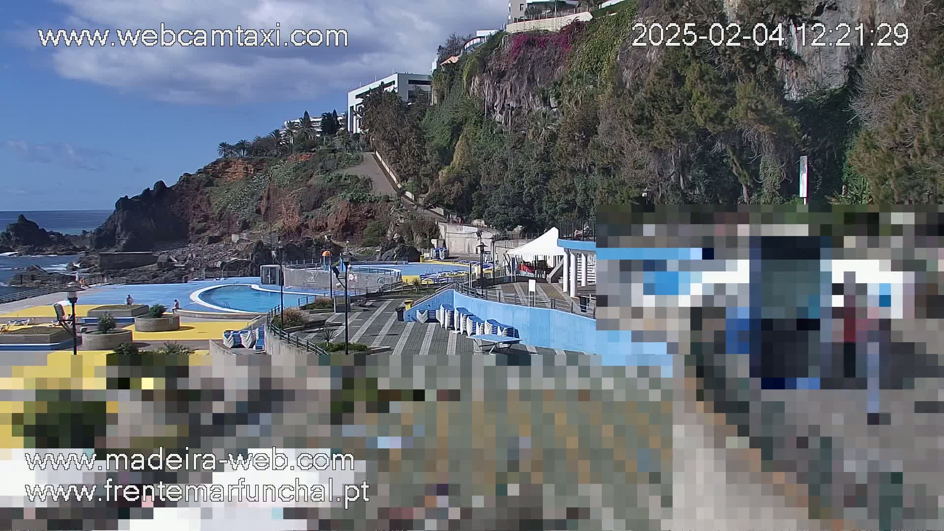 An outdoor seaside pool complex with blue tiled features is situated on a rocky cliffside under a mostly sunny sky.