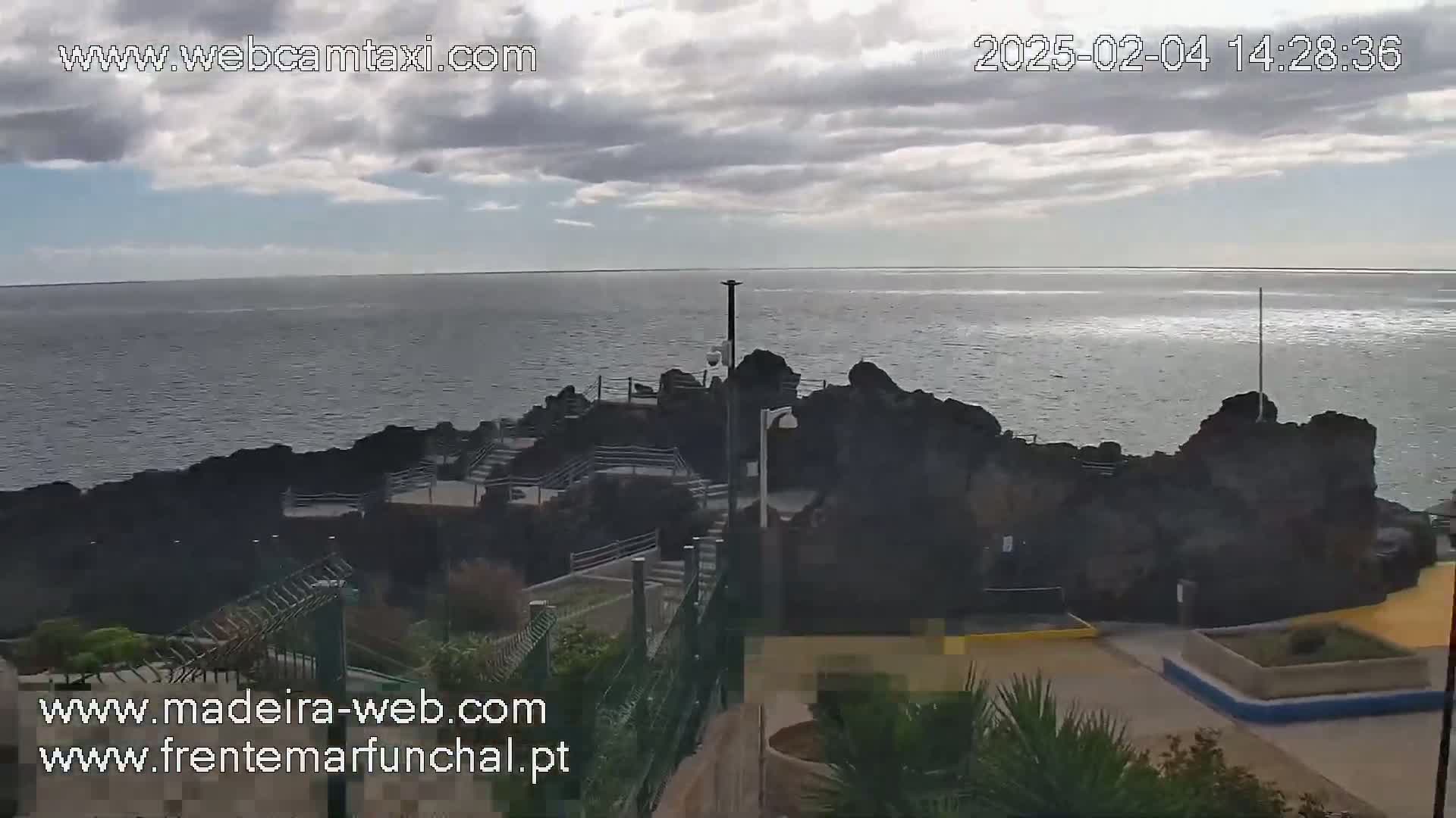 A partly cloudy day reveals a rocky coastline meeting a calm ocean, with stairs and pathways leading down to the water's edge.