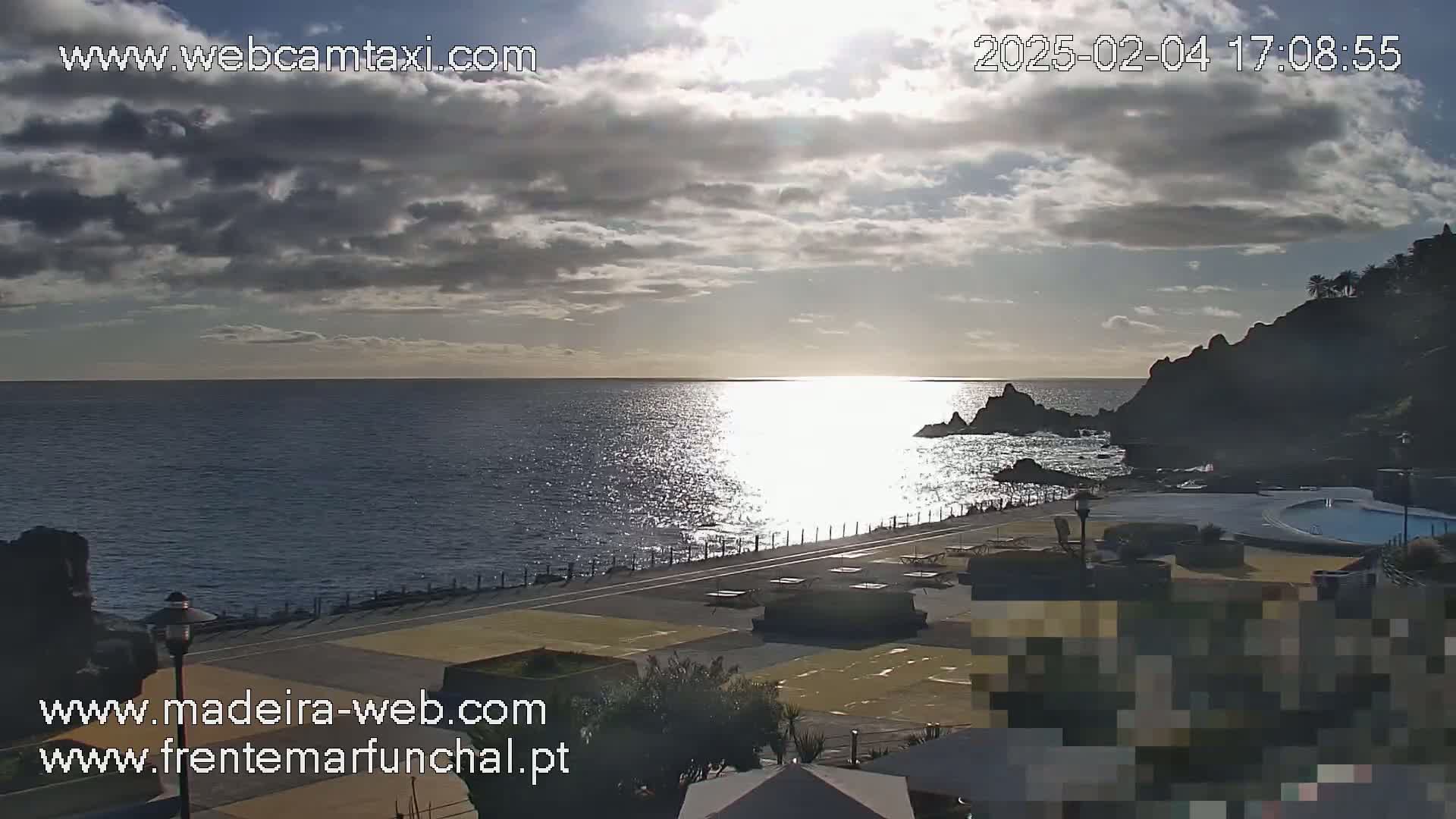 A partly cloudy sunset over a calm ocean, viewed from a coastal area with paved walkways and a swimming pool.