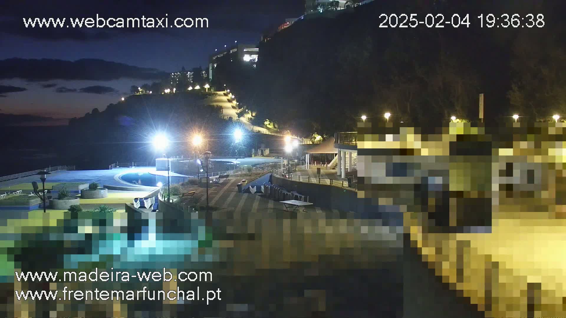 A nighttime view of a coastal area with a pool, buildings, and a path leading up a dark hillside under a partly cloudy sky.