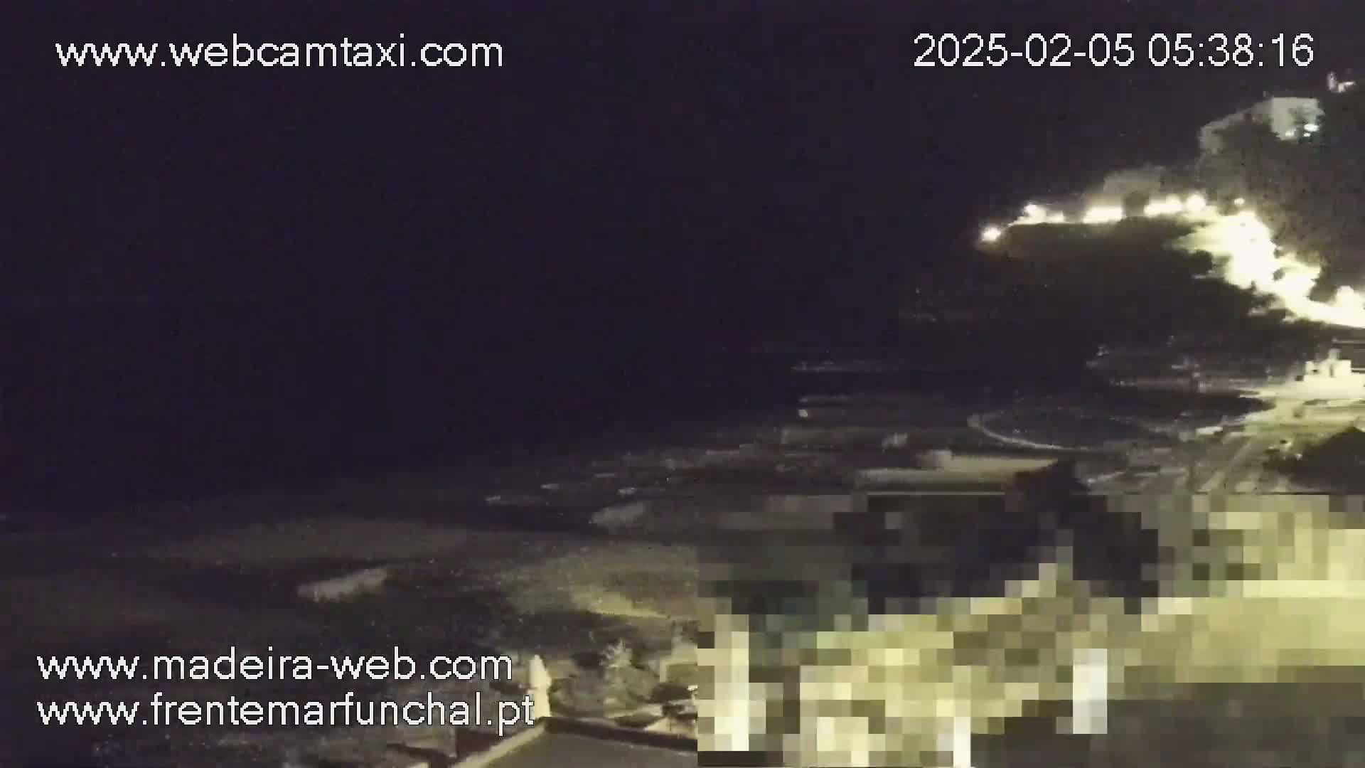 A nighttime coastal scene shows a dark ocean with waves lapping on a shore near structures with some lights visible on a hillside.