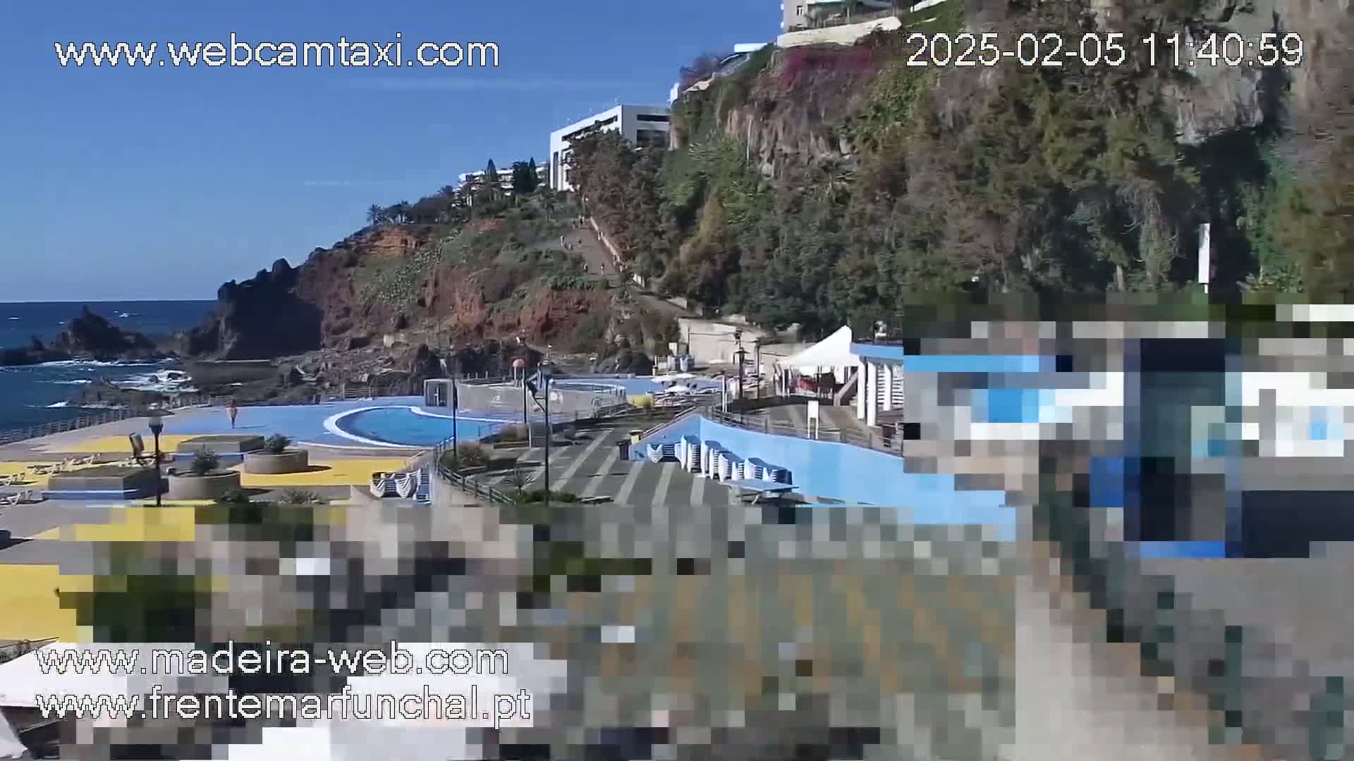 An outdoor scene on a sunny day shows an oceanside pool complex nestled against a rocky cliff, with buildings and vegetation visible on the hillside above.