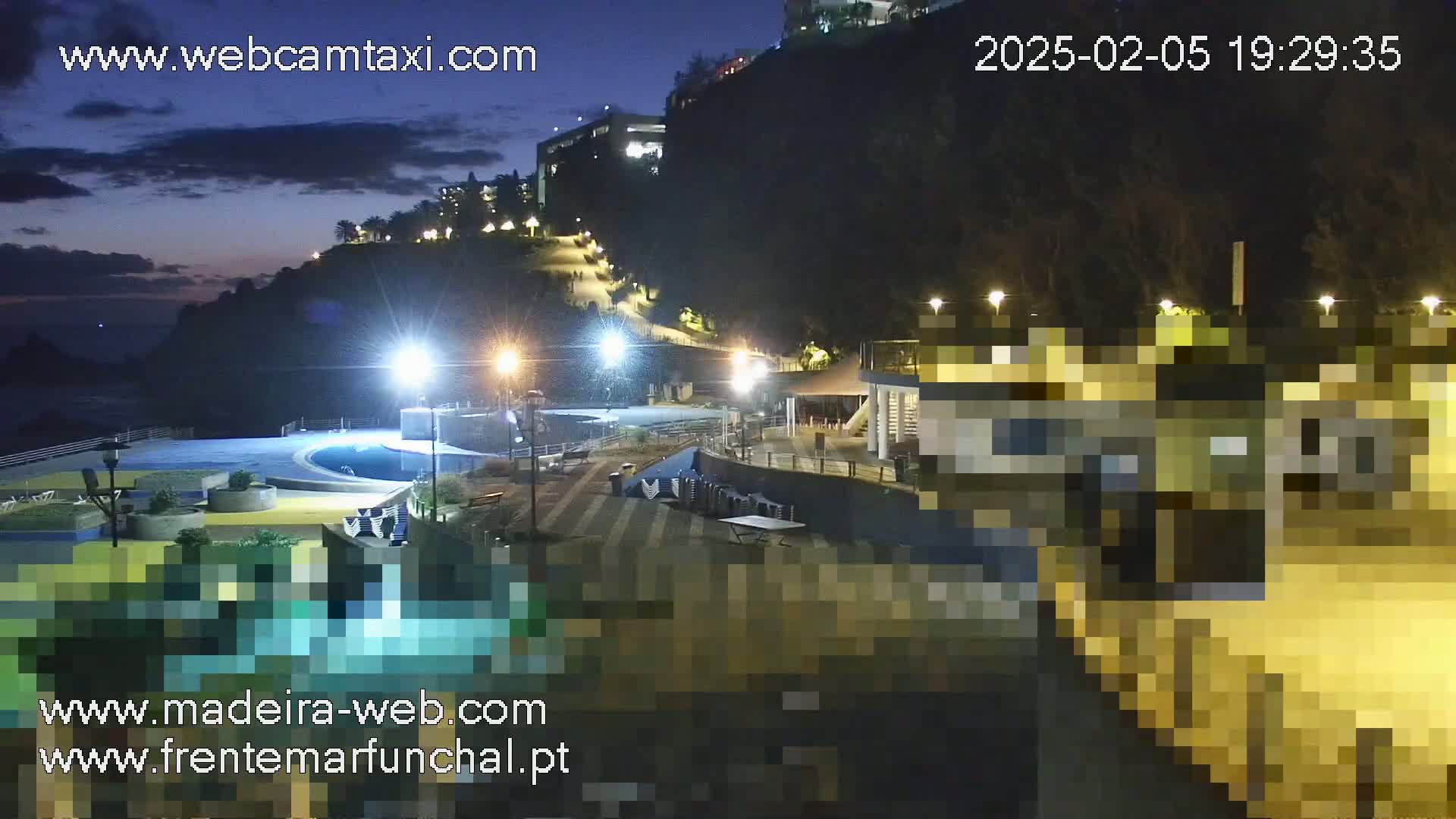 A nighttime view of a coastal area with buildings, a pool, and pathways illuminated by lights under a partly cloudy sky.