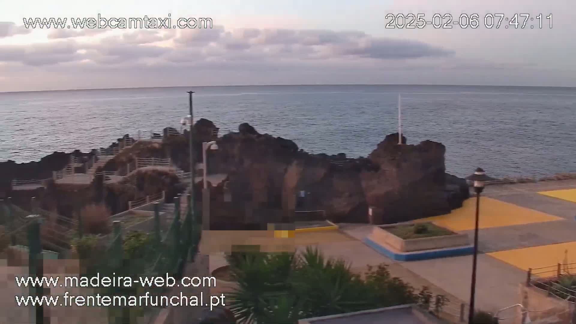 A mostly cloudy sunrise scene shows dark volcanic rock formations along a paved waterfront area overlooking a calm ocean.