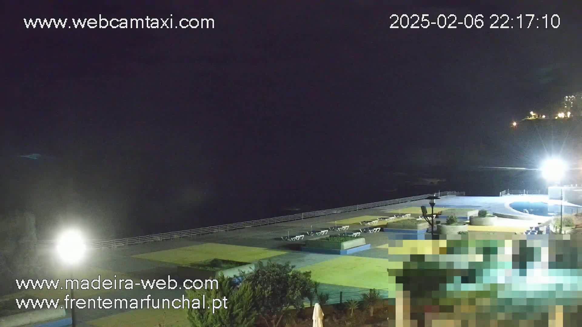 A nighttime, outdoor view of a seaside area with a pool, deck chairs, and landscaping, under a dark sky.