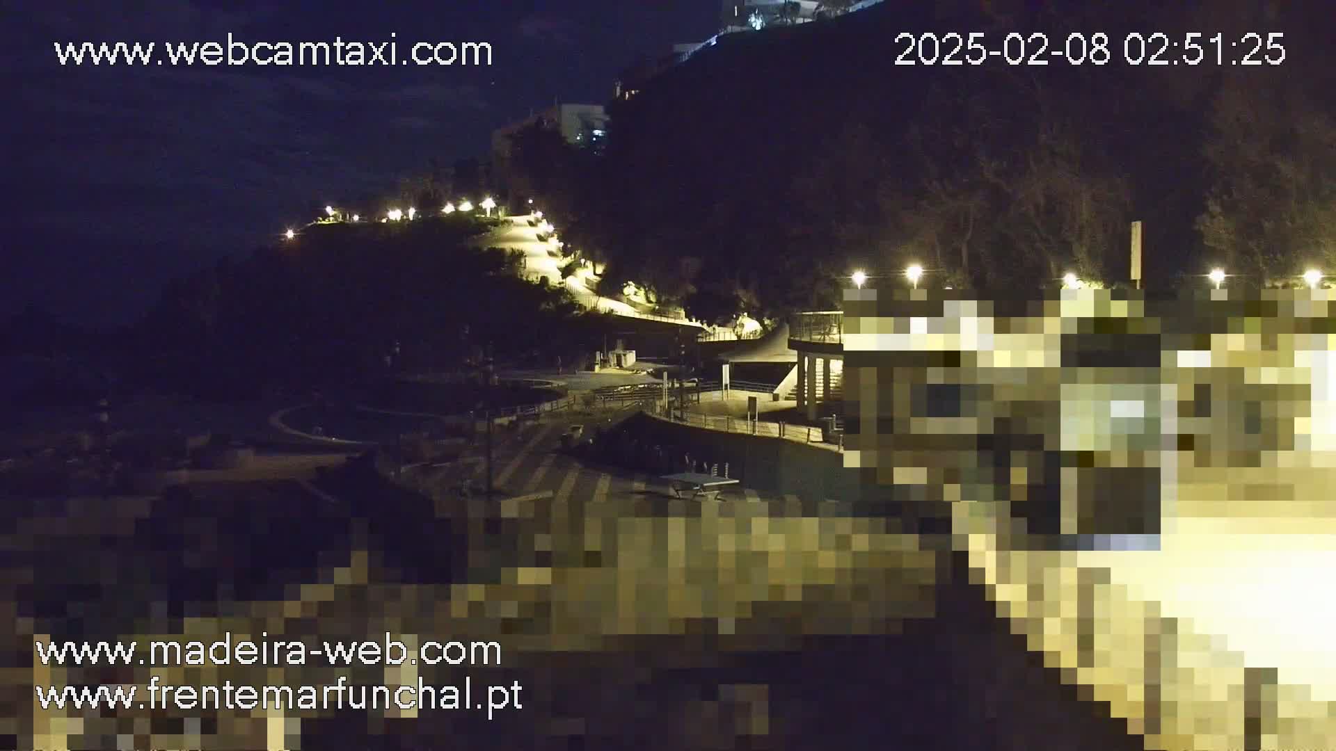 A nighttime view of a coastal area with a paved path winding up a hillside, illuminated by streetlights, leading to a building near the water.