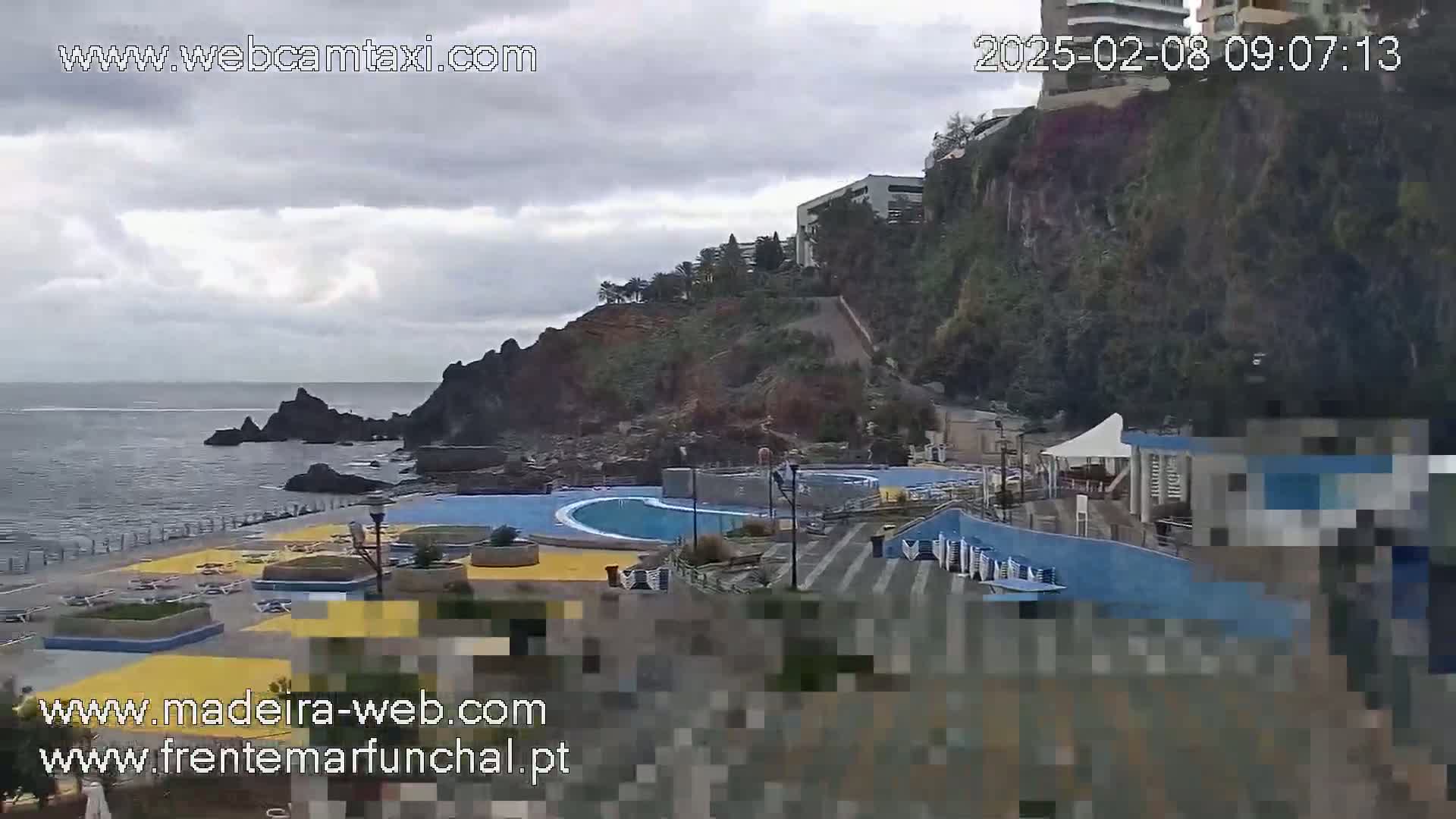 An outdoor scene on an overcast day shows a seaside swimming pool complex situated on a rocky coast next to a cliff with buildings on top.