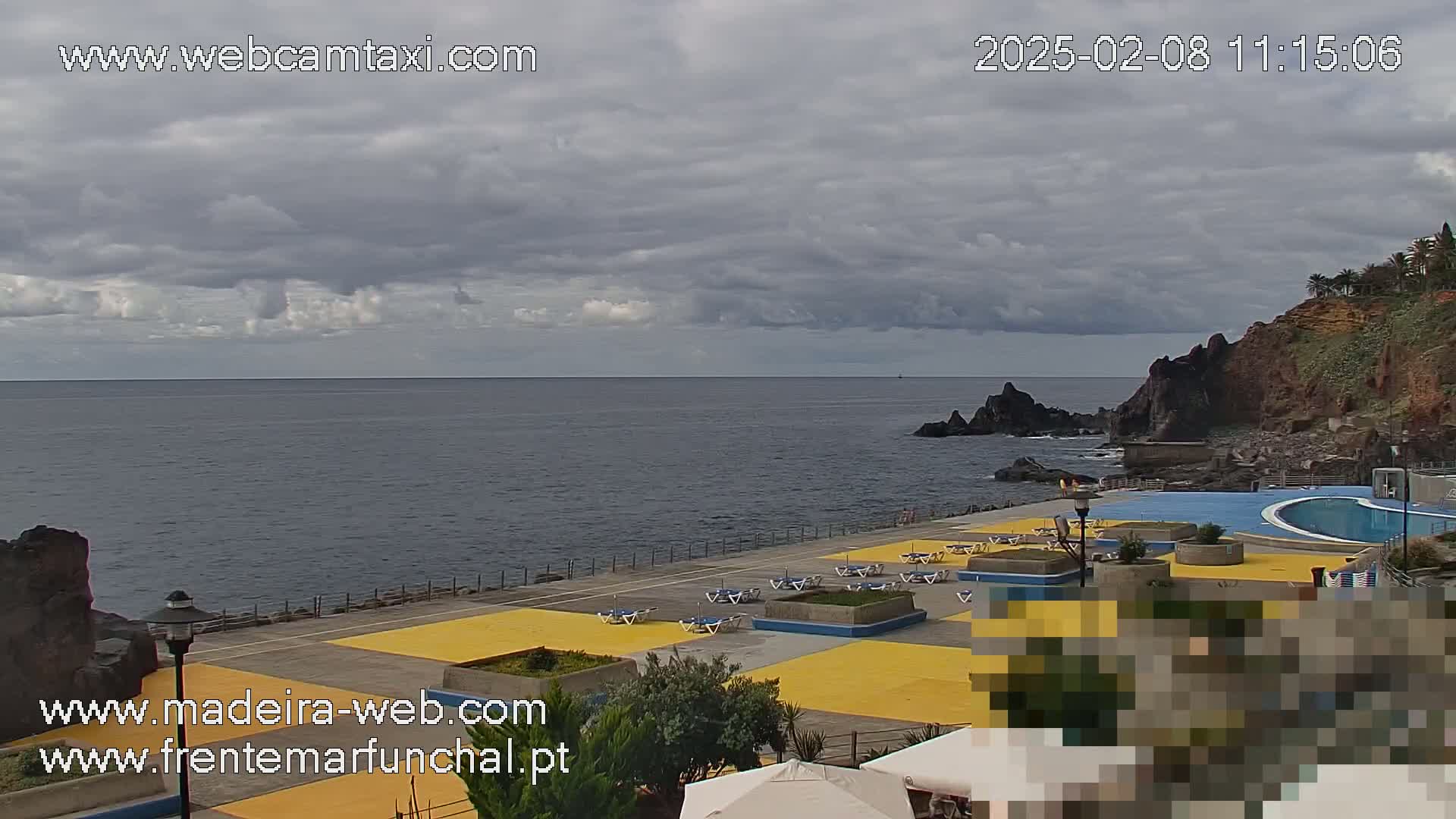 An outdoor scene shows a swimming pool and sunbathing area next to a rocky coast under a cloudy sky.