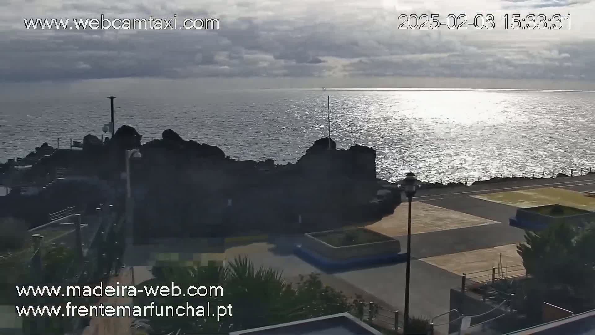 A mostly sunny view of a rocky coastline and paved waterfront area, with calm, sparkling water extending to the horizon.