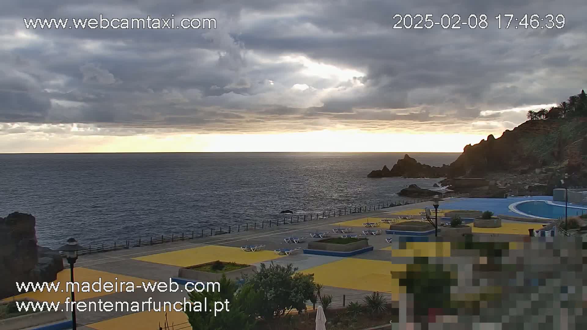 A partly cloudy sunset over a calm ocean, overlooking a seaside area with an outdoor pool and deck chairs.