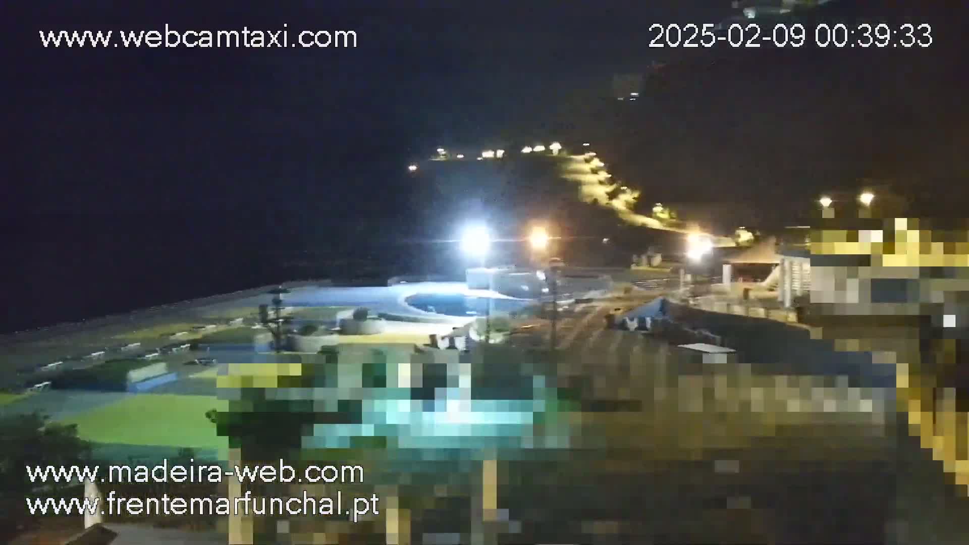 A nighttime view of a coastal area shows a pool, buildings, and a hillside lit with lights.