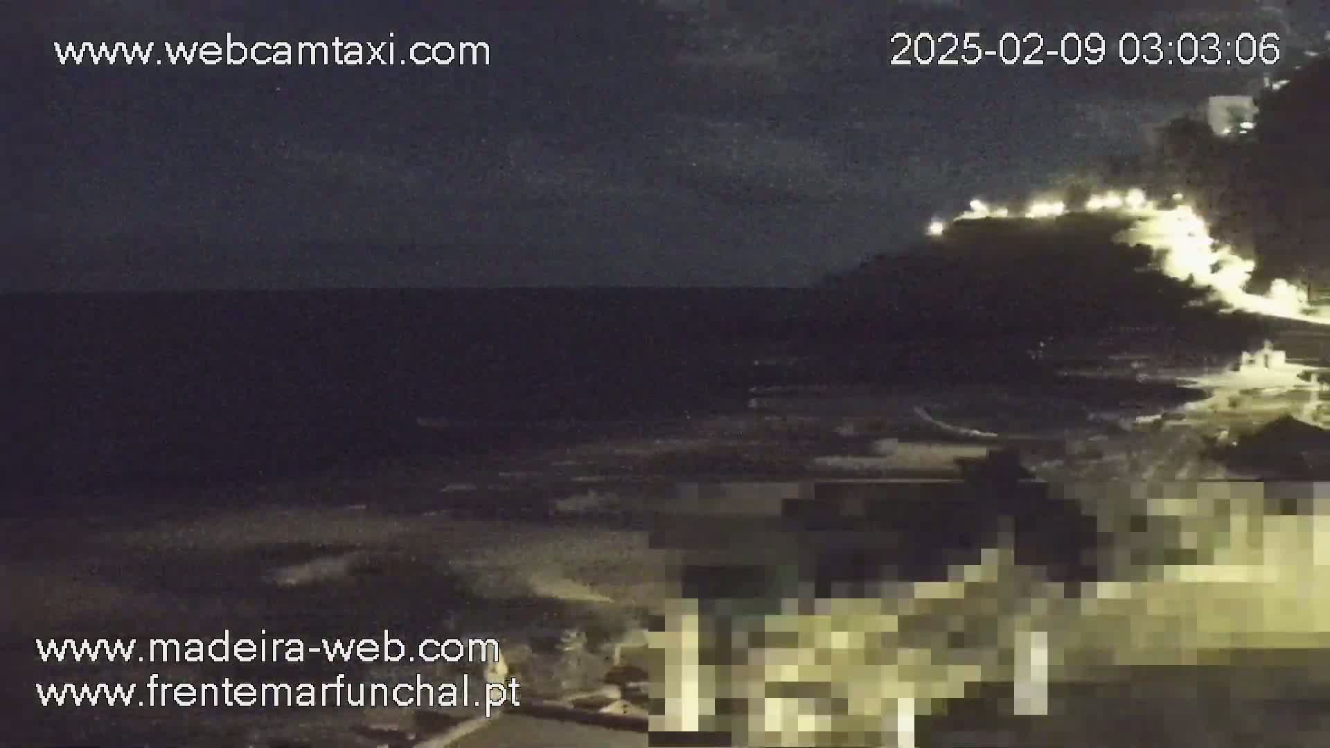 A nighttime view of a dark ocean with a hilltop illuminated by lights in the distance under a partly cloudy sky.