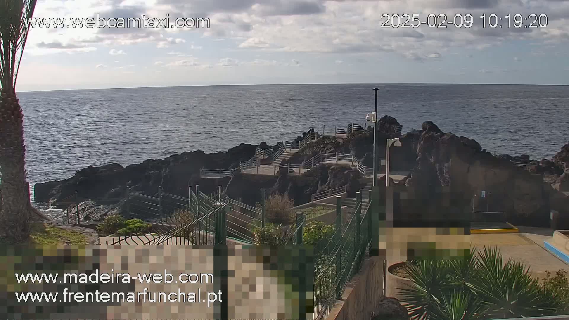 A rocky coastline with a paved walkway and railings overlooks a calm ocean under a partly cloudy sky.
